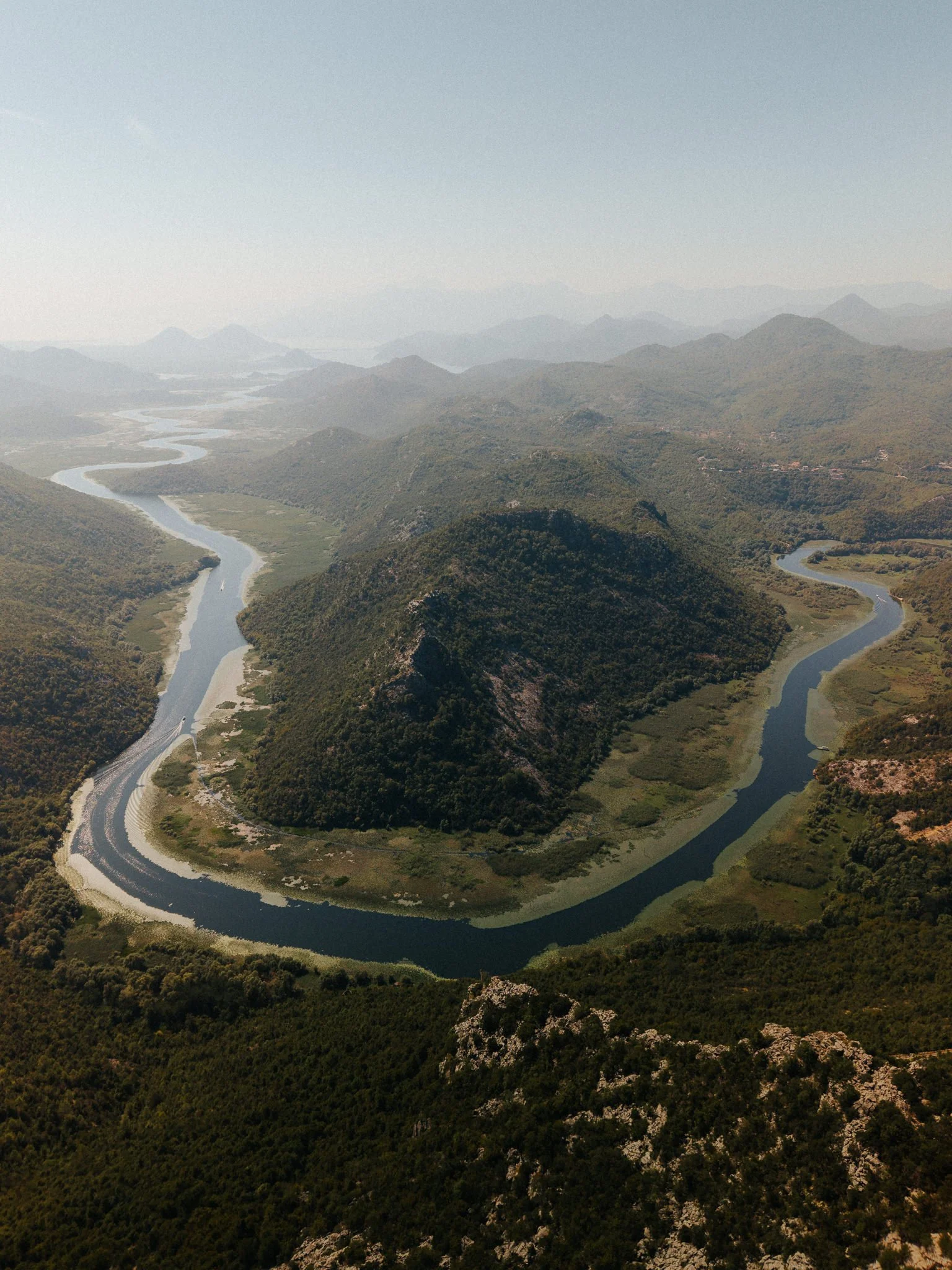 Aerial view of a winding river flowing through green, forested mountains under a hazy sky.