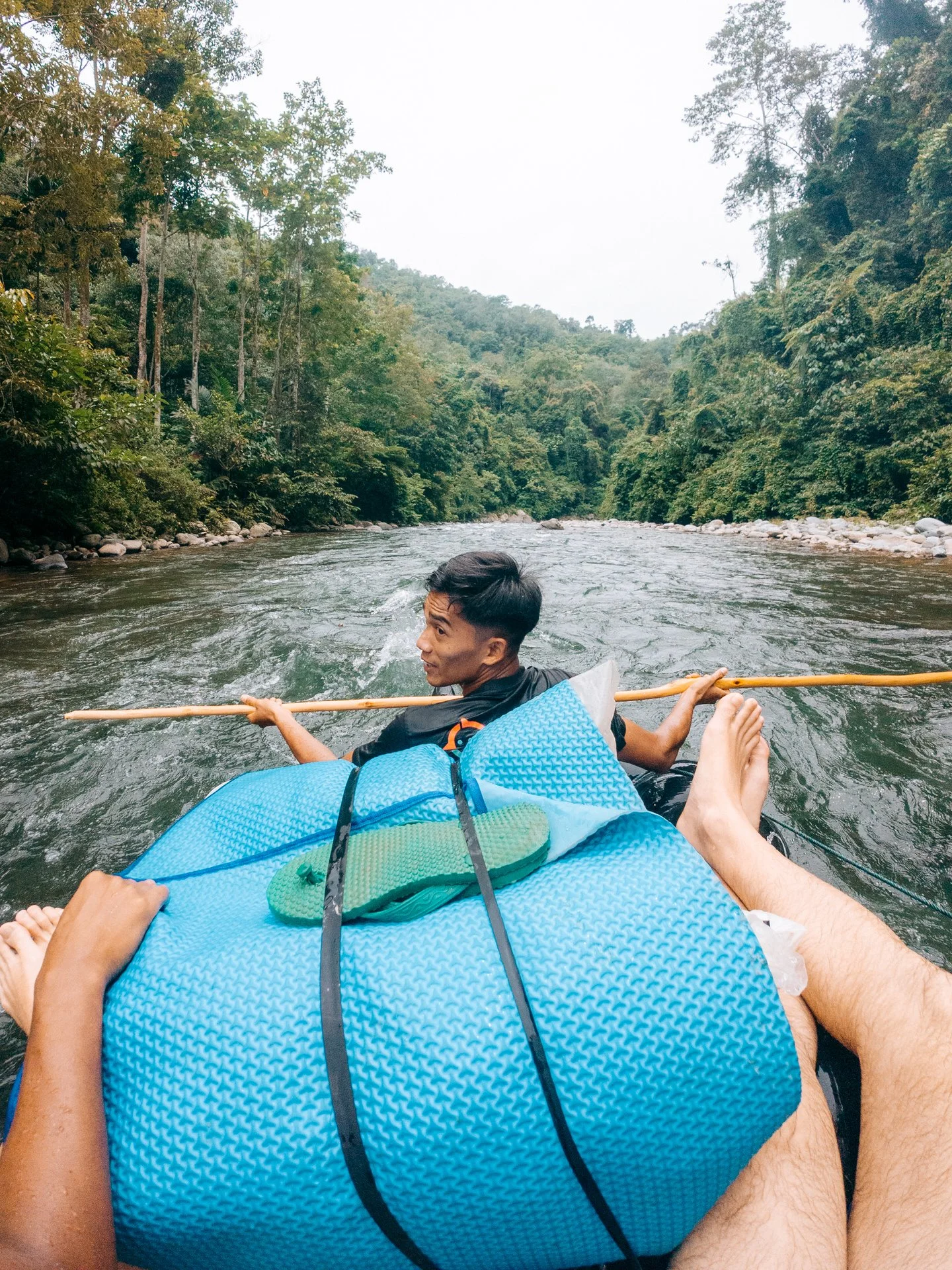 Person floating on a river in a blue raft with another person ahead holding a stick, surrounded by lush green trees and hills.