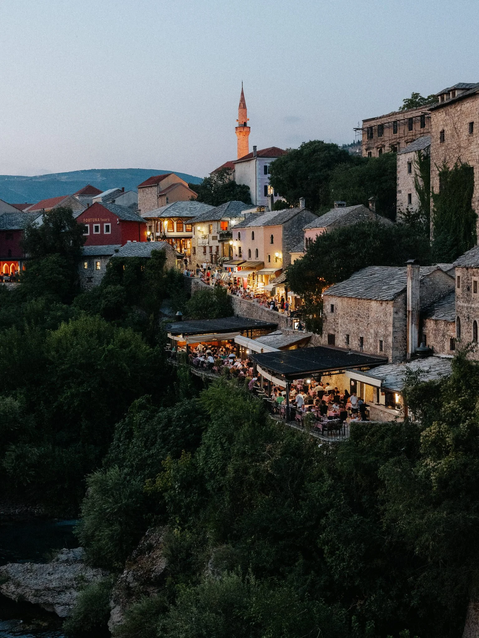 A scenic view of a historic European town at dusk with a river in the foreground. The town has stone buildings with red-tiled roofs, a minaret, and a lively street filled with outdoor cafes and people dining under string lights.