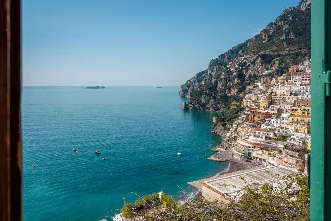 View of a seaside village built on a cliff, with colorful houses stacked on the hillside and boats floating in the calm blue water, seen through a window frame.
