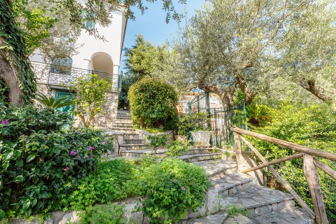 Stone steps leading up to a white house with arched doorways, surrounded by lush green plants and trees on a sunny day.