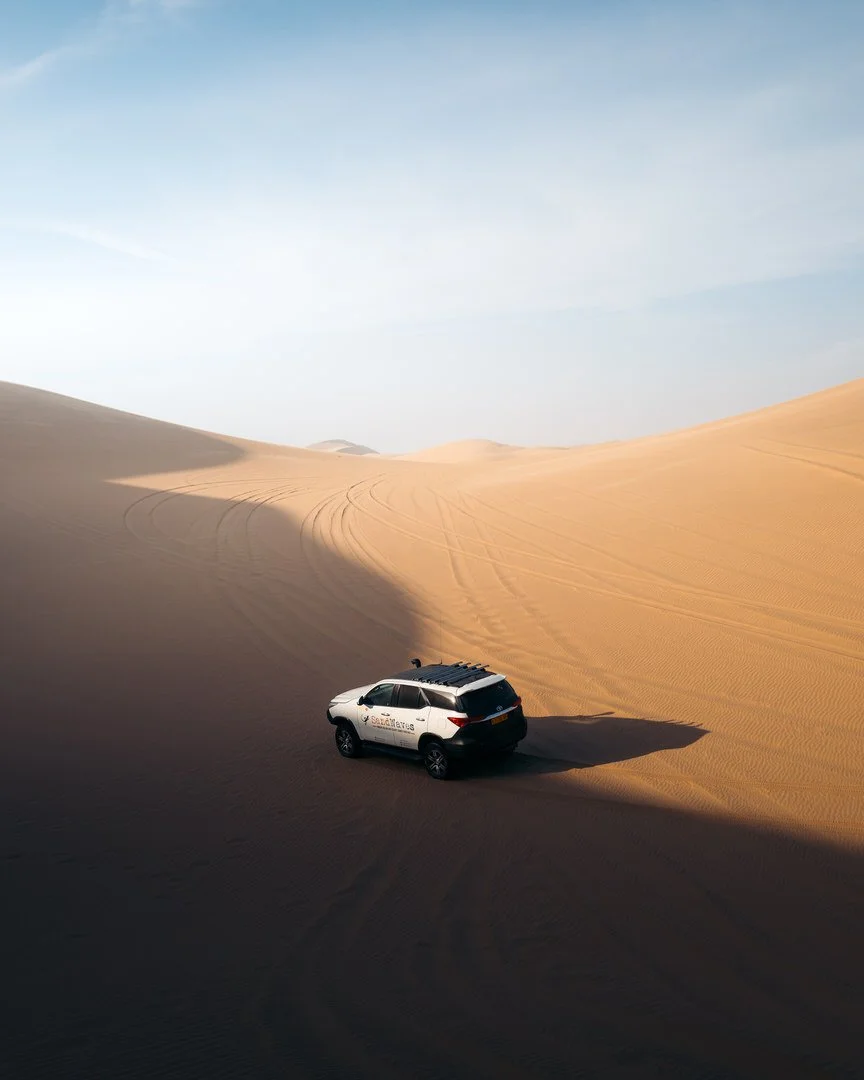 A white SUV driving through a desert with tall sand dunes and tire tracks, under a clear sky.