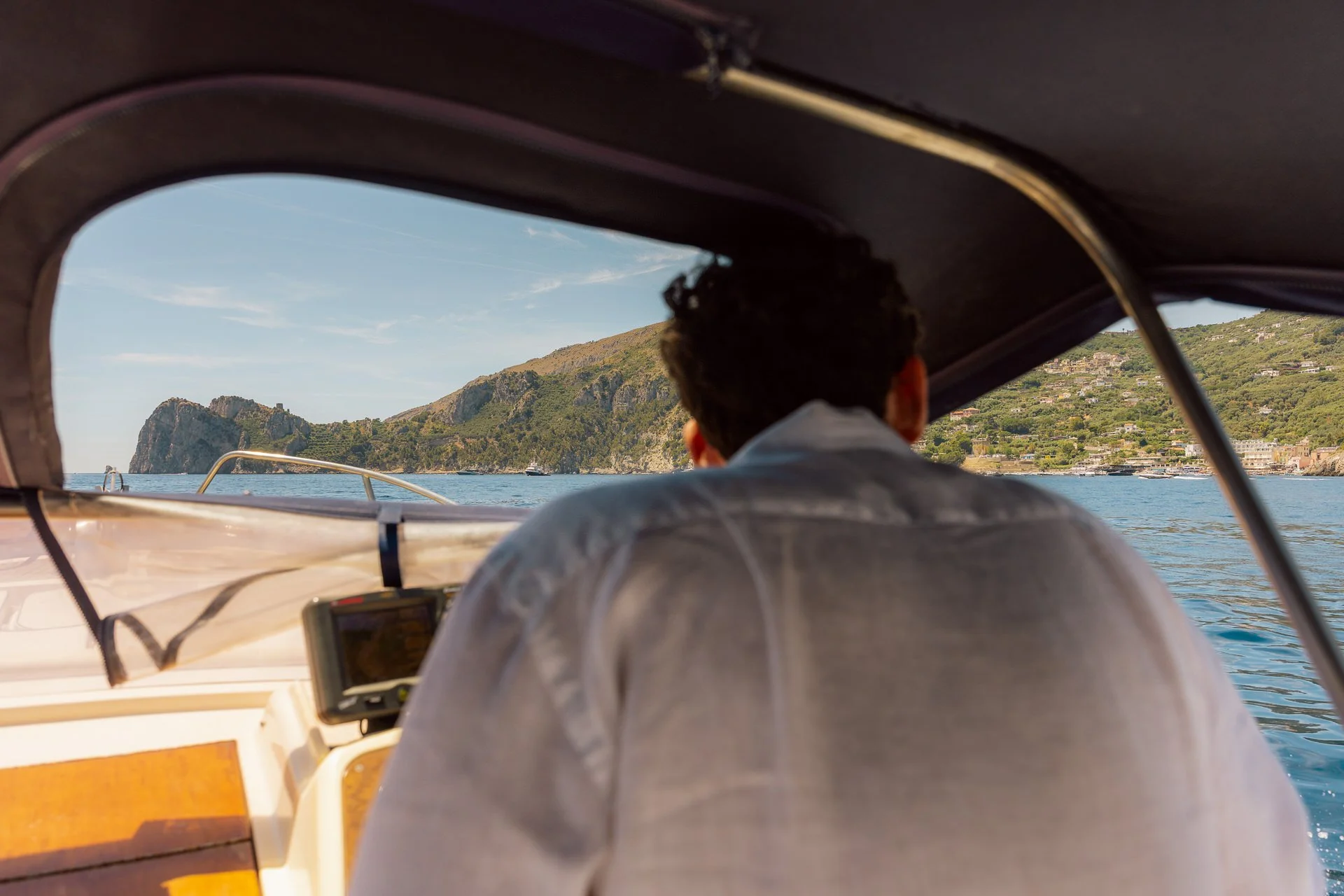 A person with dark hair, wearing a white or light-colored shirt, is sitting on a boat, looking out at the water and hills in the distance, with a coastline and clear blue sky.