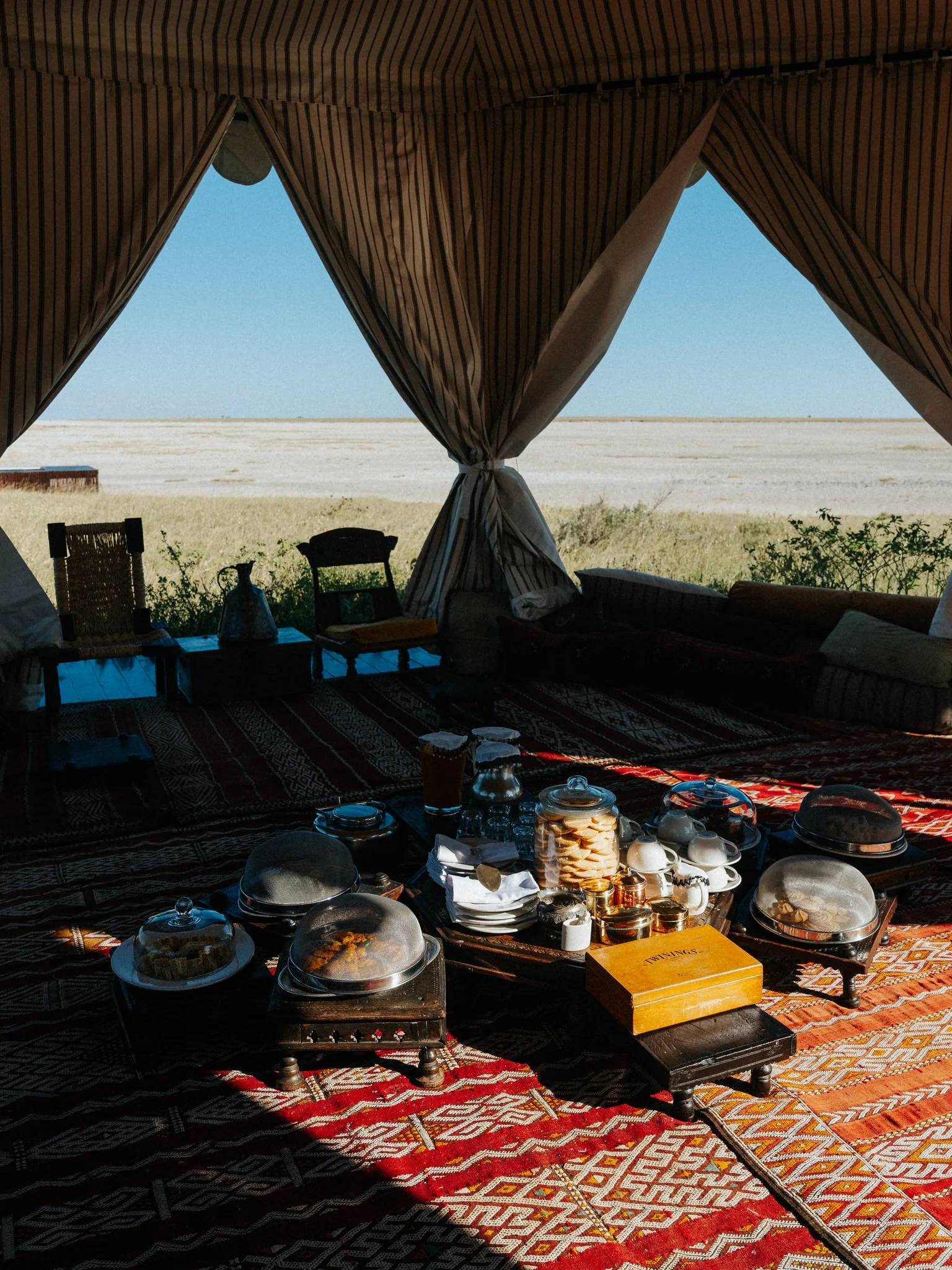 View of an outdoor desert landscape with a clear blue sky, seen through the open doorway of a tent. Inside, there is a cozy seating area with cushions, a table with tea and snacks, and various dishes and glassware.