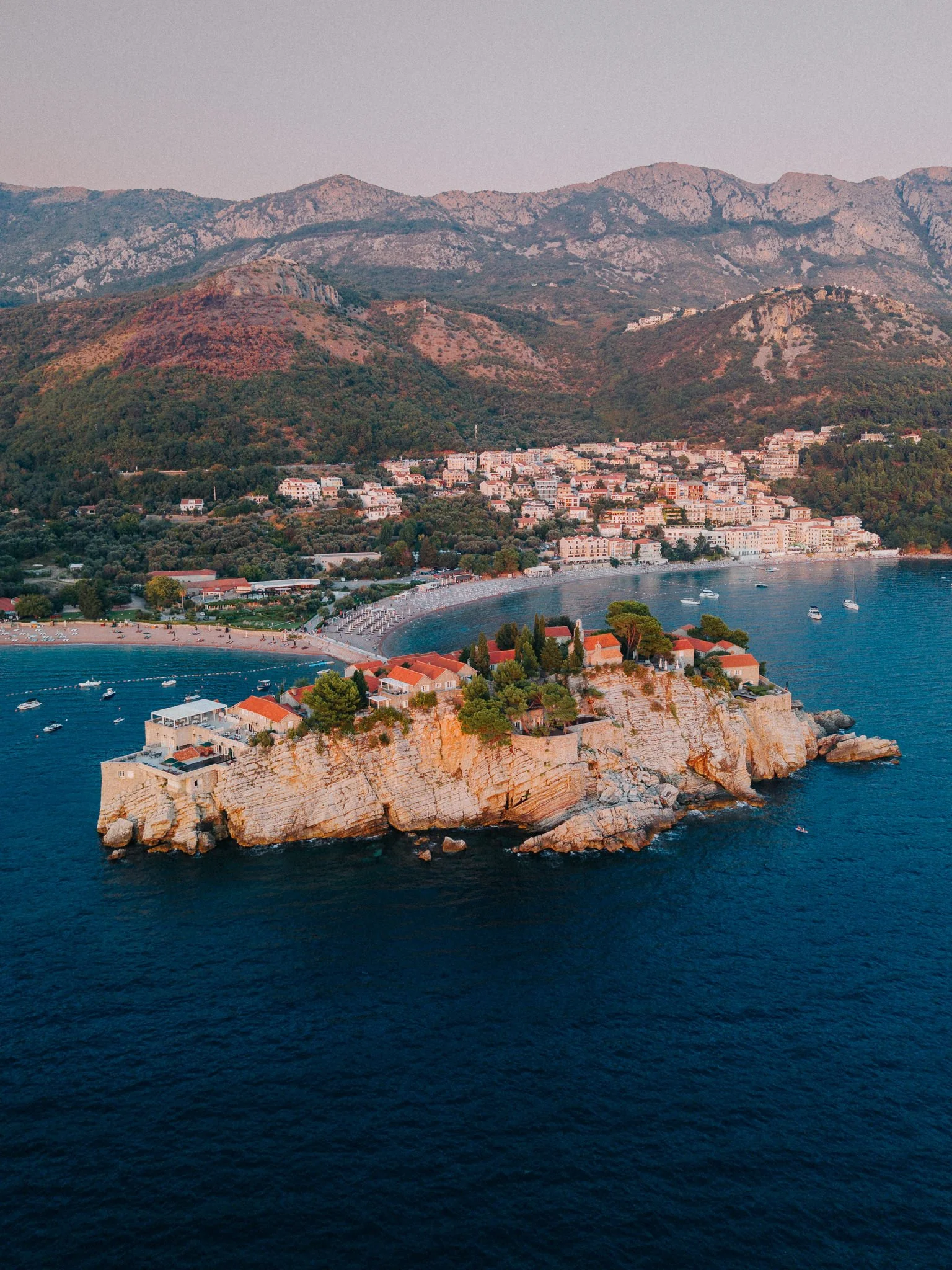 Coastal town with a rocky island in the foreground, beach, and colorful hillside with buildings and green trees, surrounded by deep blue water.