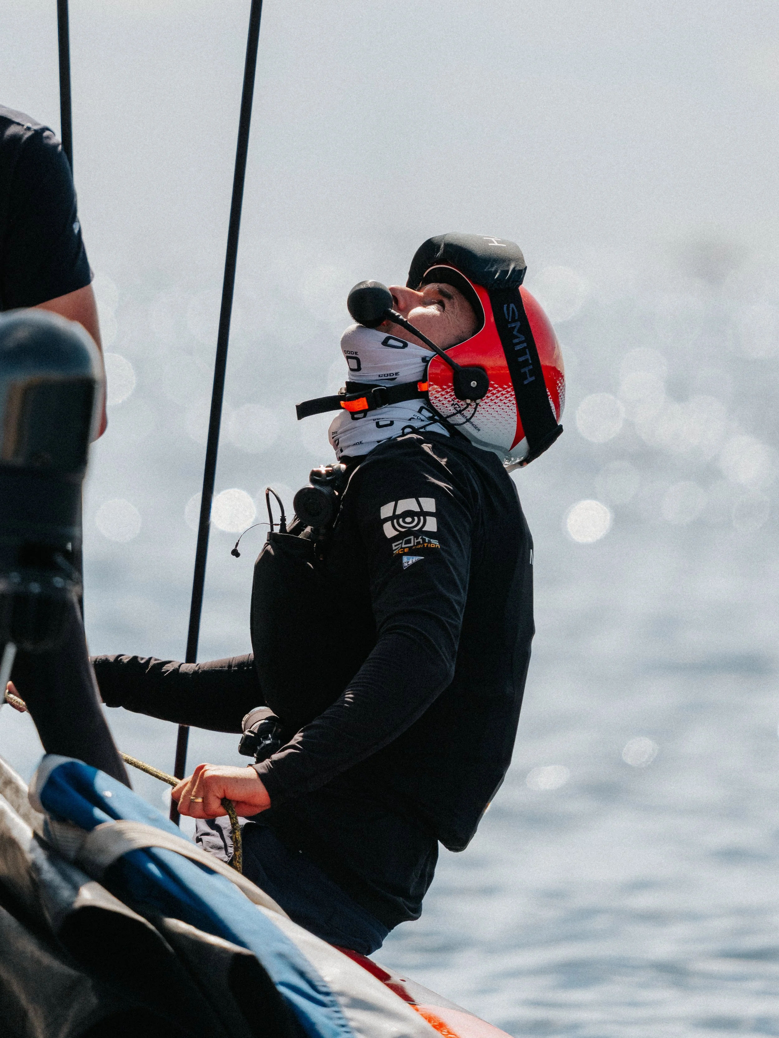A person in a racing helmet and racing gear, sitting in a kayak, adjusting or holding a paddle, with water and cloudy sky in the background.