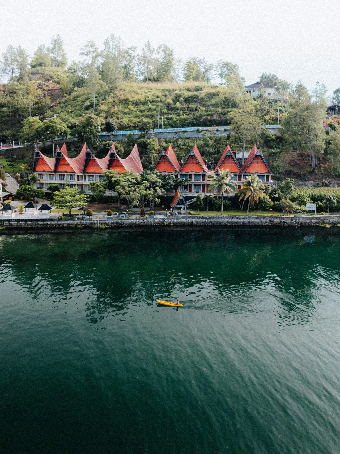 A scenic lakeside view featuring a row of traditional styled buildings with red pointed roofs, lush green trees, and a hill in the background. There is a small boat with a person paddling on the calm water in the foreground.