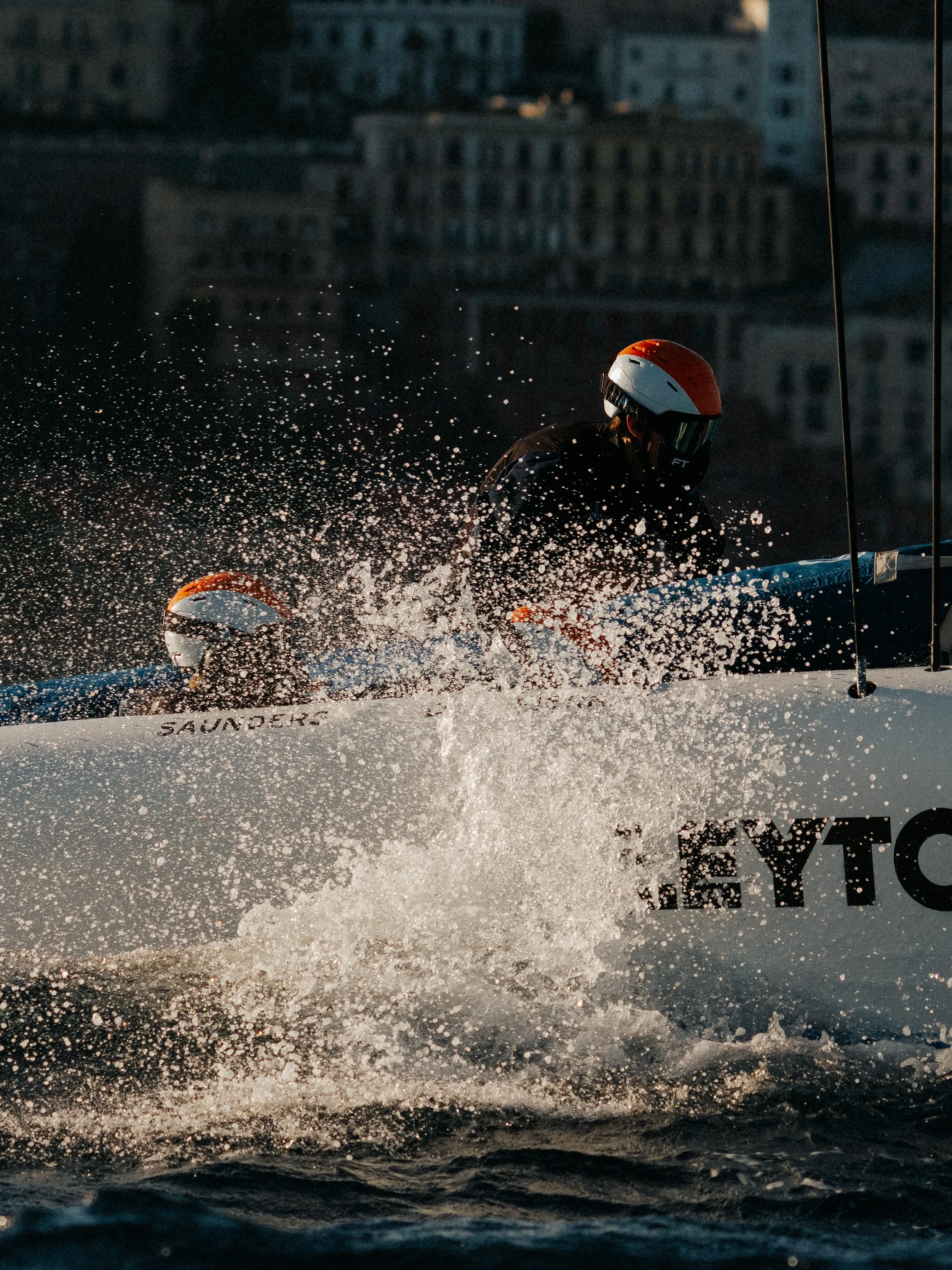 An emergency responder or cyclist wearing a helmet navigating through water spray or splashes, possibly during a race or training, with a cityscape in the background.