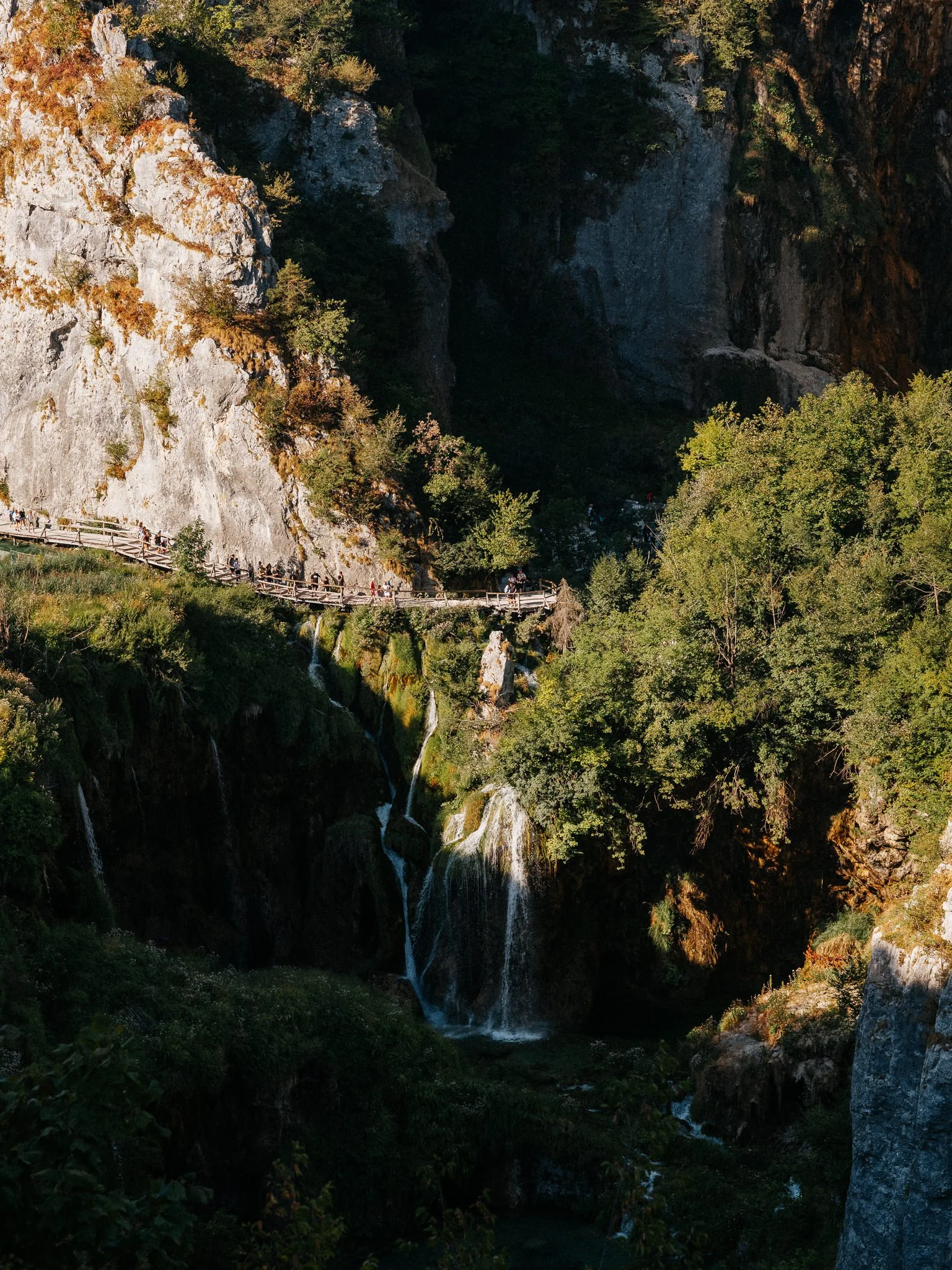 A rocky landscape with a tall cliff and a small waterfall, lush green trees, and a wooden boardwalk with people walking on it.