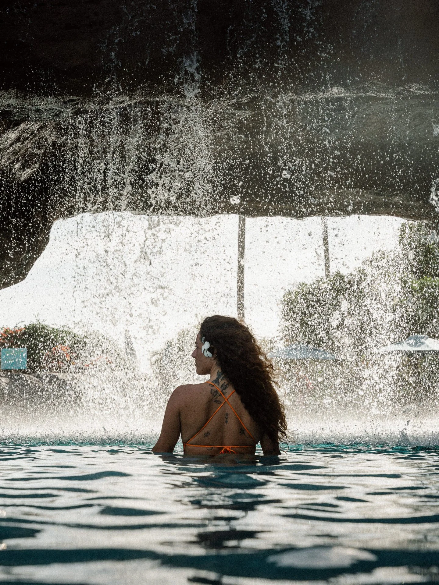 A woman with curly hair, wearing a swimsuit and headphones with a flower, stands in the water under a waterfall, facing sideways.
