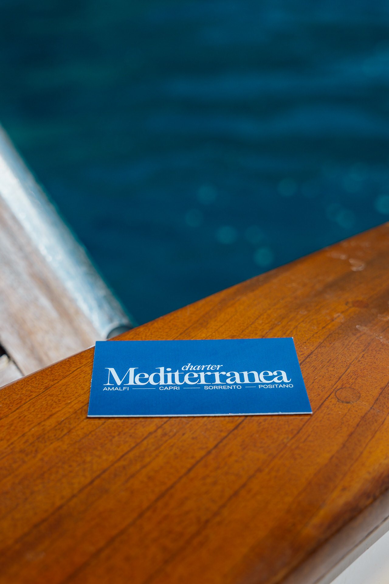 Blue business card with white text reading 'charter Mediterranea' and city names 'Amalfi, Capri, Sorrento, Positano' placed on a wooden surface near a body of water.