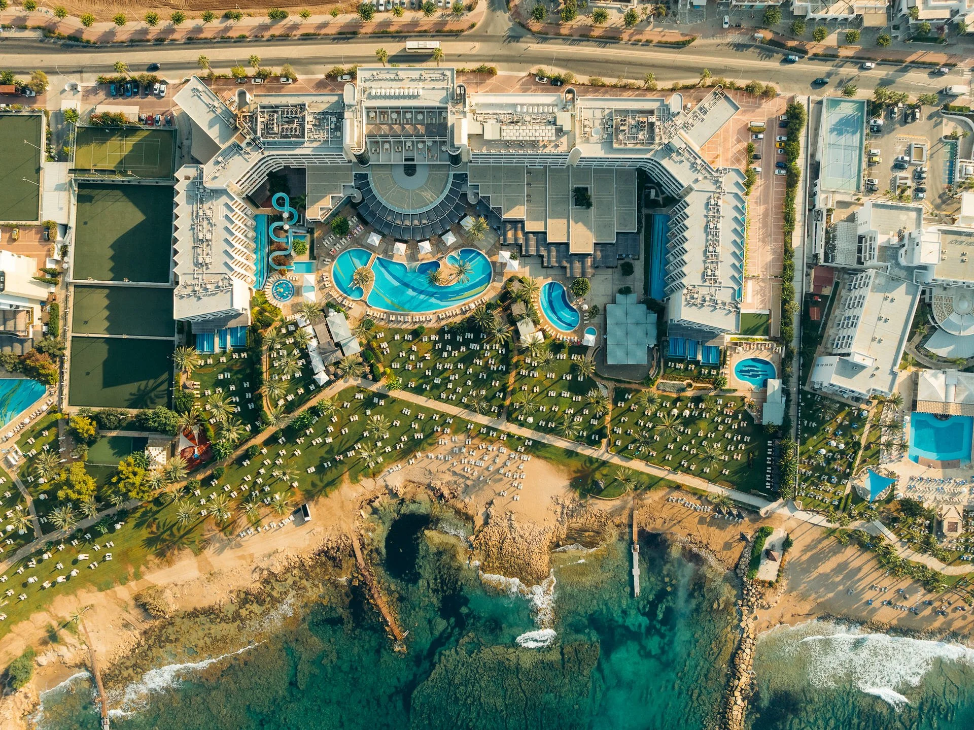Aerial view of a beachfront hotel with a large pool area, surrounded by lounge chairs, palm trees, and greenery. The hotel is adjacent to the ocean with sandy beaches.