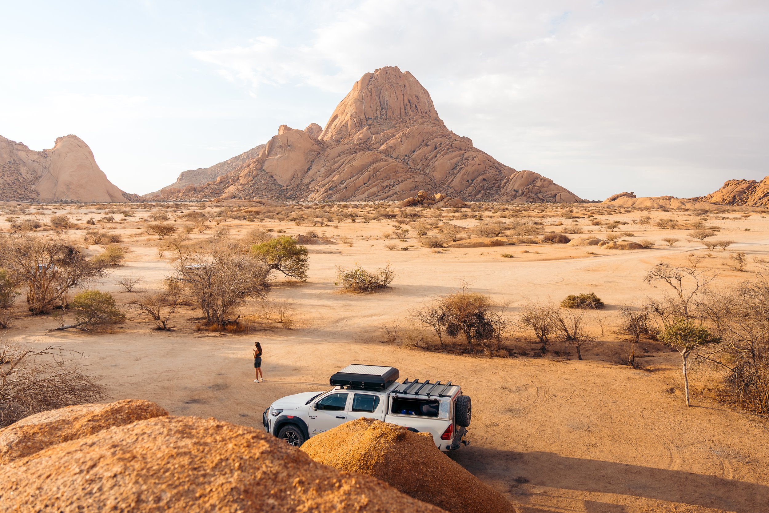 A person standing next to a white SUV in a desert landscape with sparse trees, rocky terrain, and a large mountain in the background under a cloudy sky.