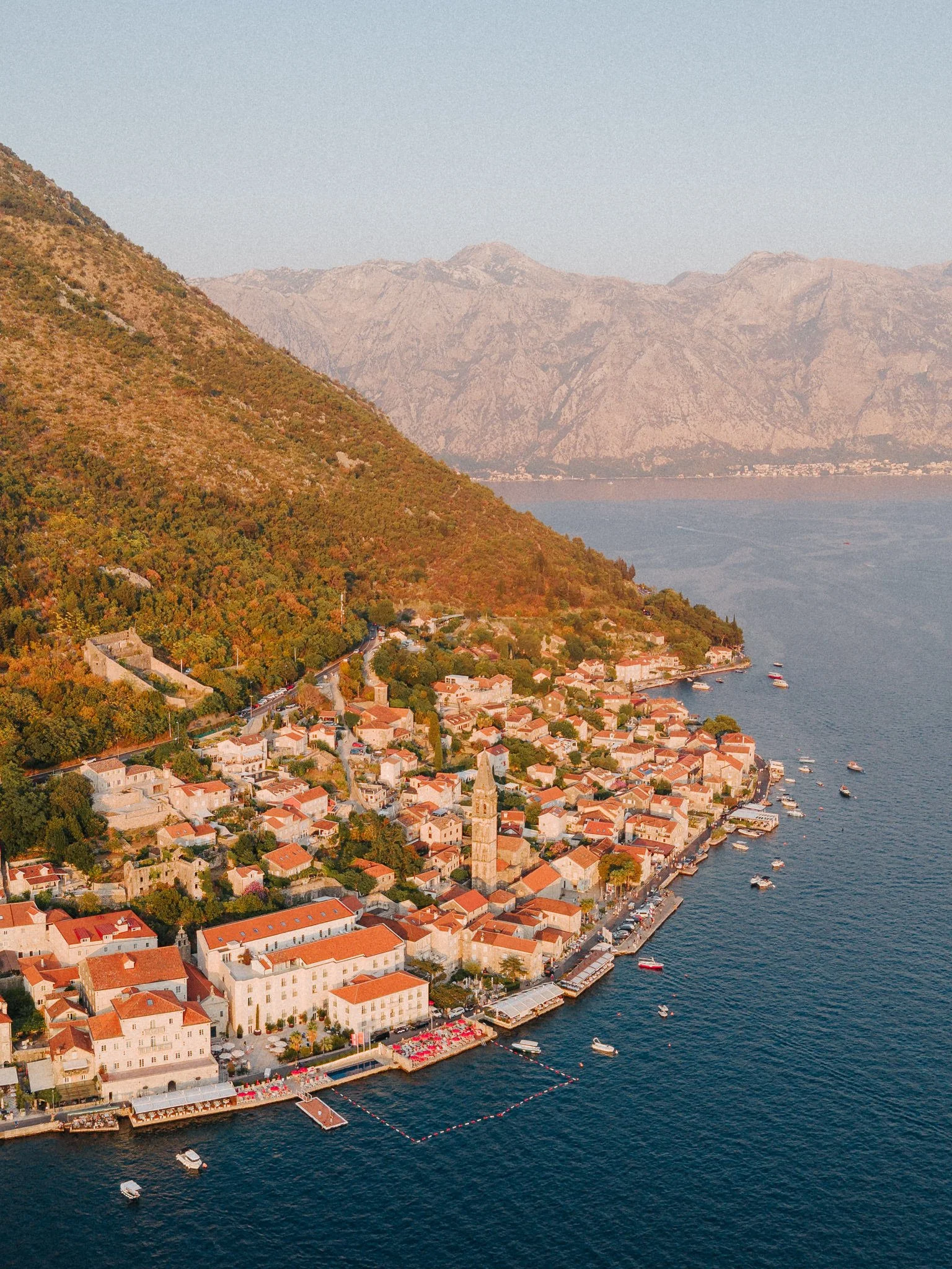 A coastal town with white buildings and red-tiled roofs along the Adriatic Sea, with a hillside covered in trees and mountains in the background.