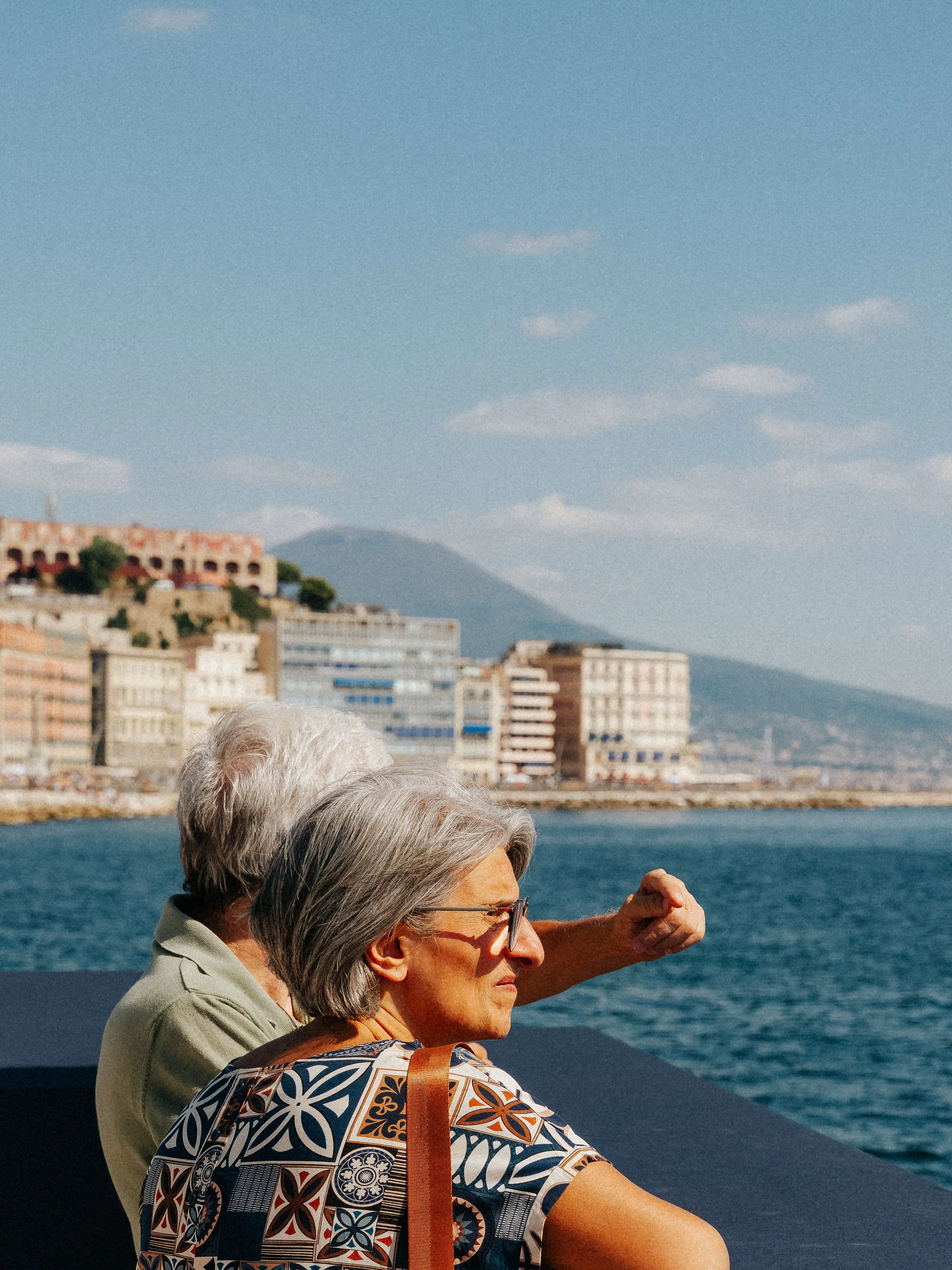 Two elderly women with gray hair and glasses sitting by the water, enjoying a sunny day with a cityscape and mountains in the background.