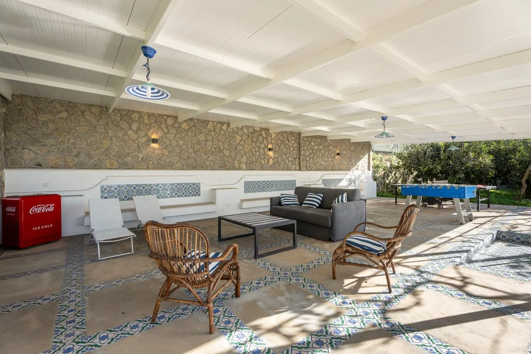 Covered outdoor patio with stone wall, white ceiling with beams, and outdoor furniture including a cushioned sofa, wicker chairs, lounge chairs, a black coffee table, and a blue foosball table on patterned tile flooring.