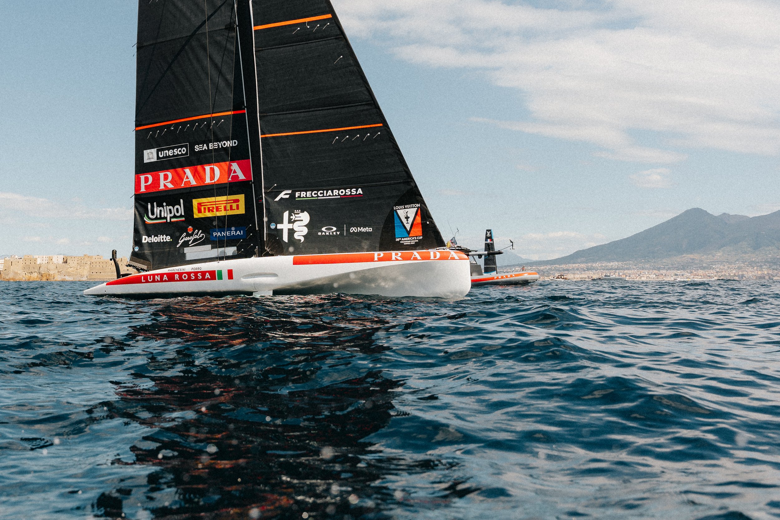 A sailboat with a black sail decorated with sponsor logos, including Prada, on the water. The boat is near a coastal city with mountains in the background under a partly cloudy sky.