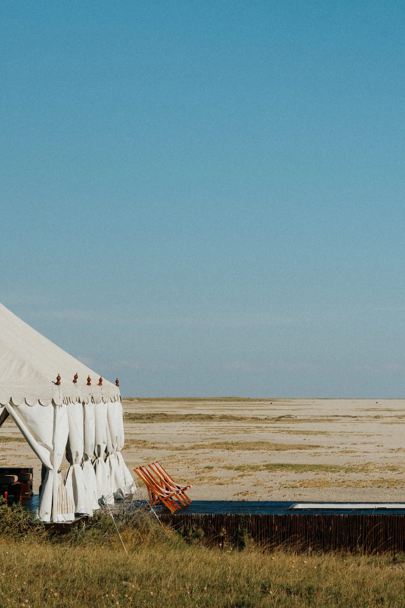 A large white tent with wooden finials on top in an open desert landscape under a clear blue sky. There are striped chairs near the tent and some beach chairs on a blue surface in the foreground, with sparse vegetation and flat terrain extending into