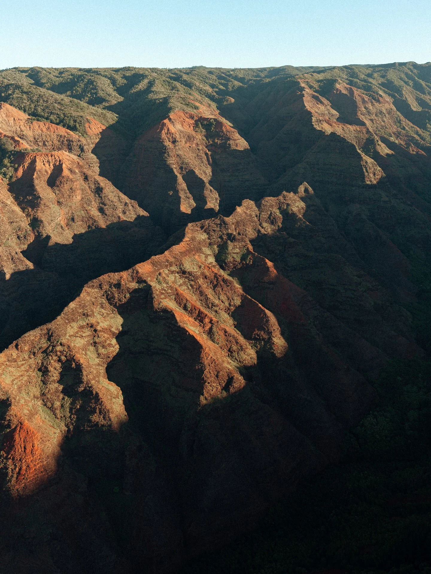 Sunlit view of the rugged, multicolored ridges of the Grand Canyon, showing shadows cast by the sunlight over the layered rock formations.