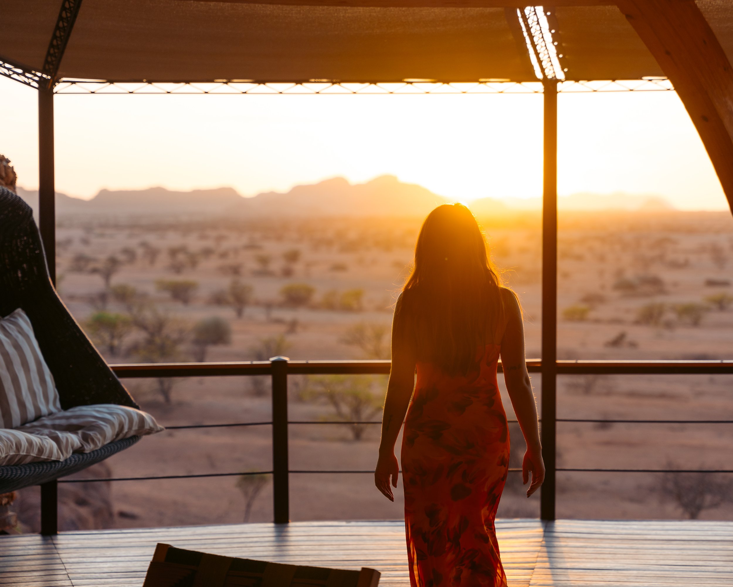 A woman in a long dress stands on a balcony watching the sunset over a desert landscape with sparse trees and mountains in the distance.
