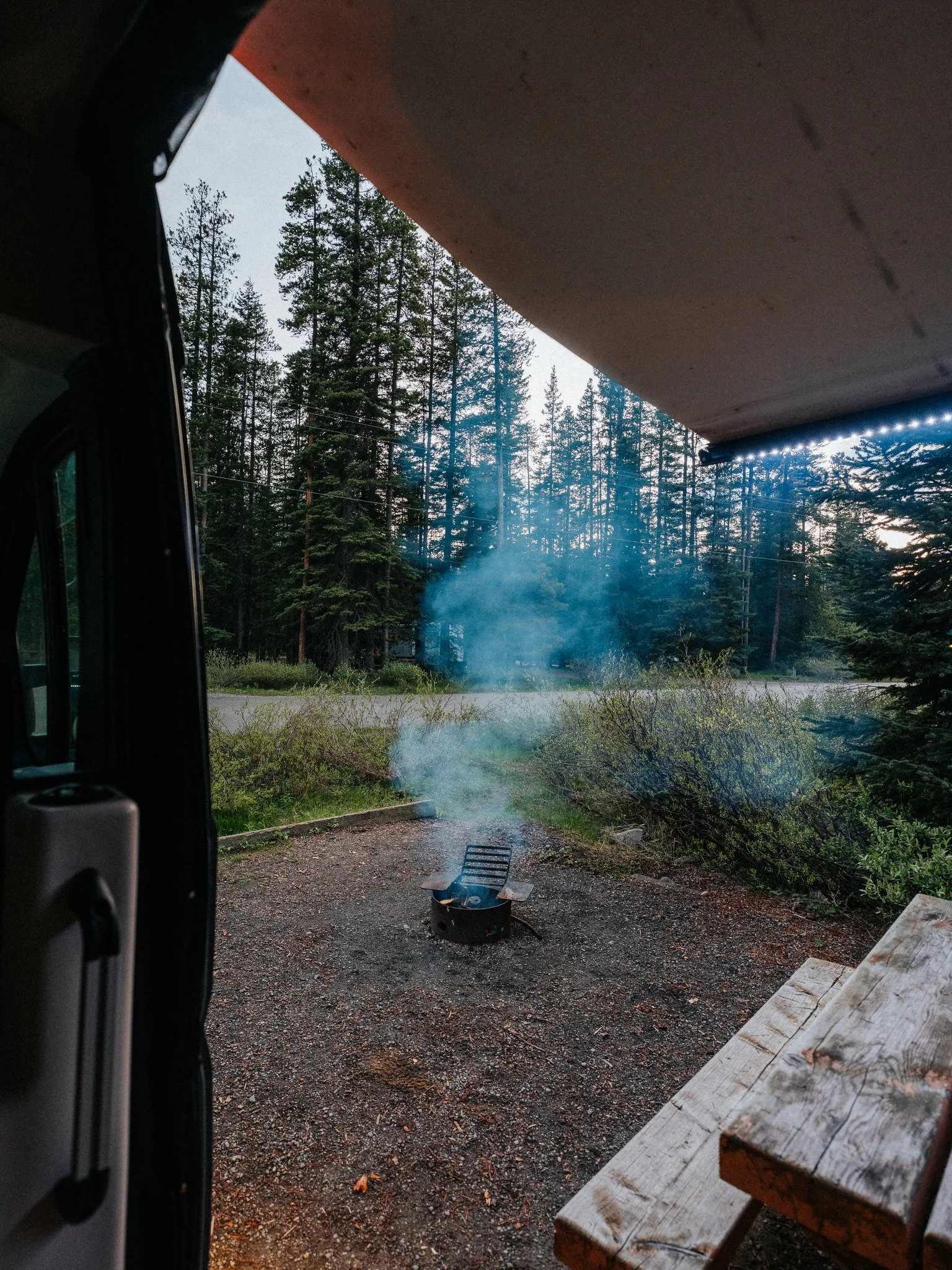 Looking out from underneath a roof overhang at a small campfire on a gravel ground, with trees and a cloudy sky in the background.