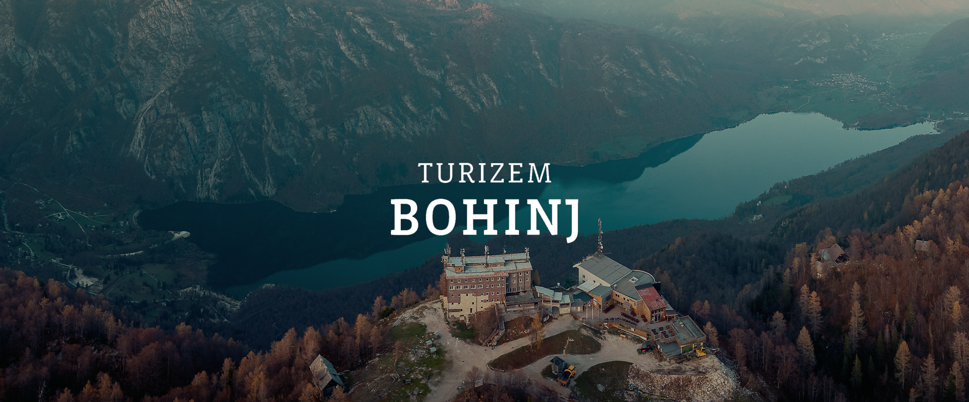 Aerial view of Lake Bohinj surrounded by mountains with a mountain lodge and a few small buildings in the foreground, overlaid with text reading "Turizem Bohinj."