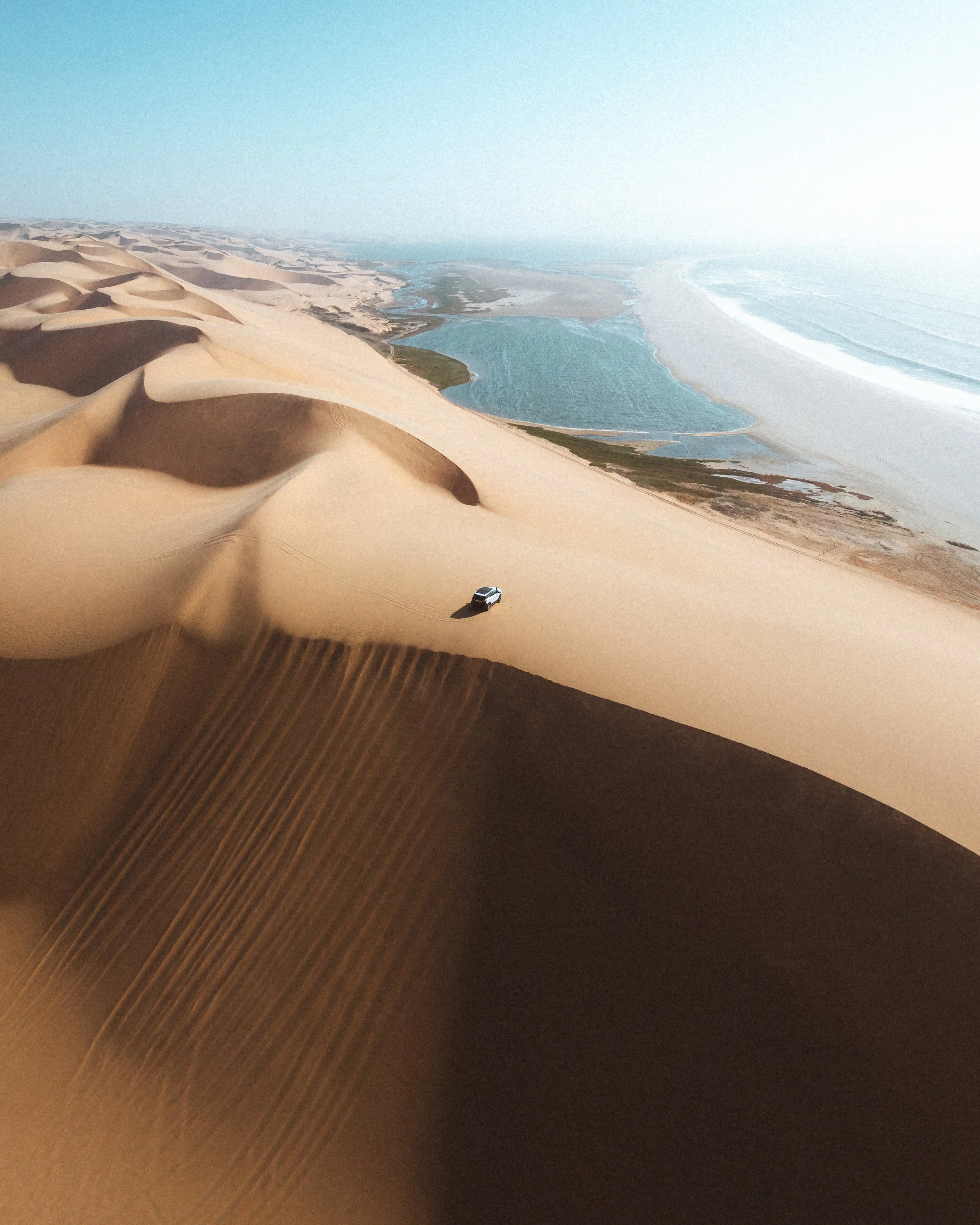 Aerial view of sand dunes in a desert with a vehicle driving on the sand and a landscape of lakes and ocean in the background.