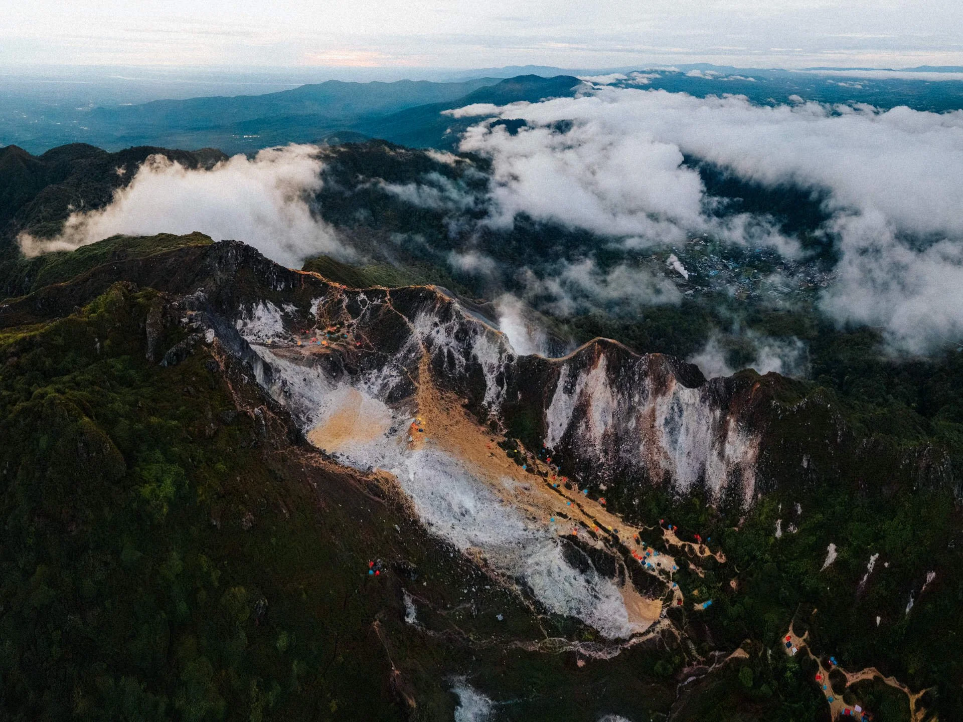 Aerial view of a mountain with landslide debris, tents, and a winding path, surrounded by lush green forests and cloud-covered mountain peaks.