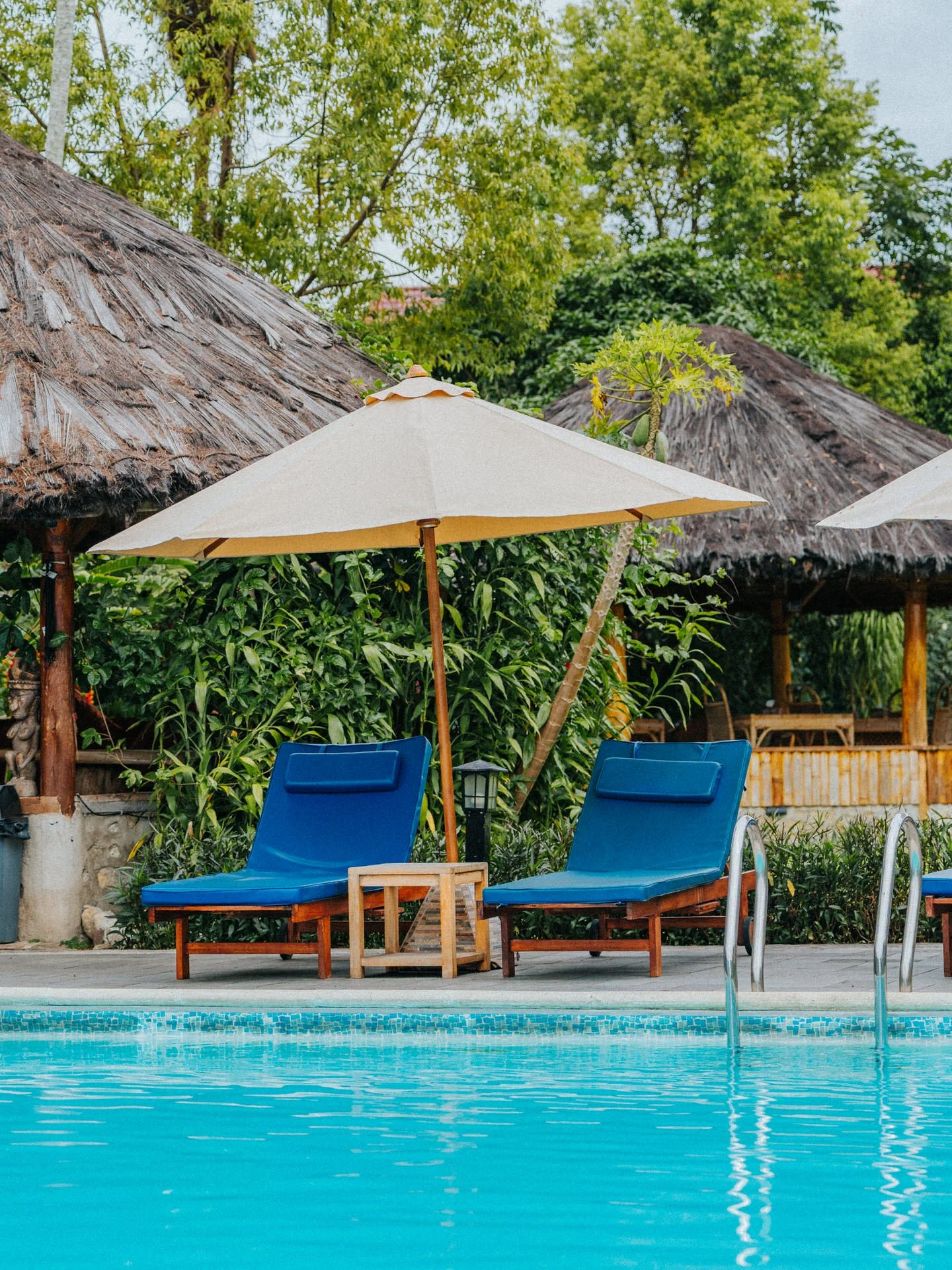 Poolside scene with two blue lounge chairs, white umbrellas, lush green trees, and thatched-roof structures in the background.