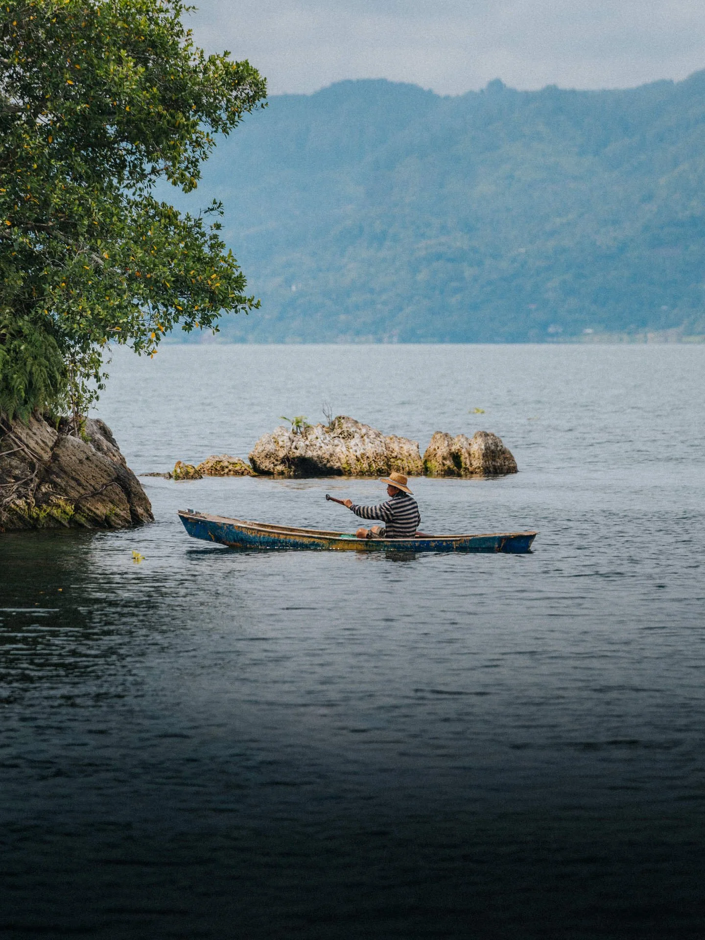 A person in a striped shirt and straw hat paddling a small boat near a rocky shoreline with lush greenery, with hills in the background.