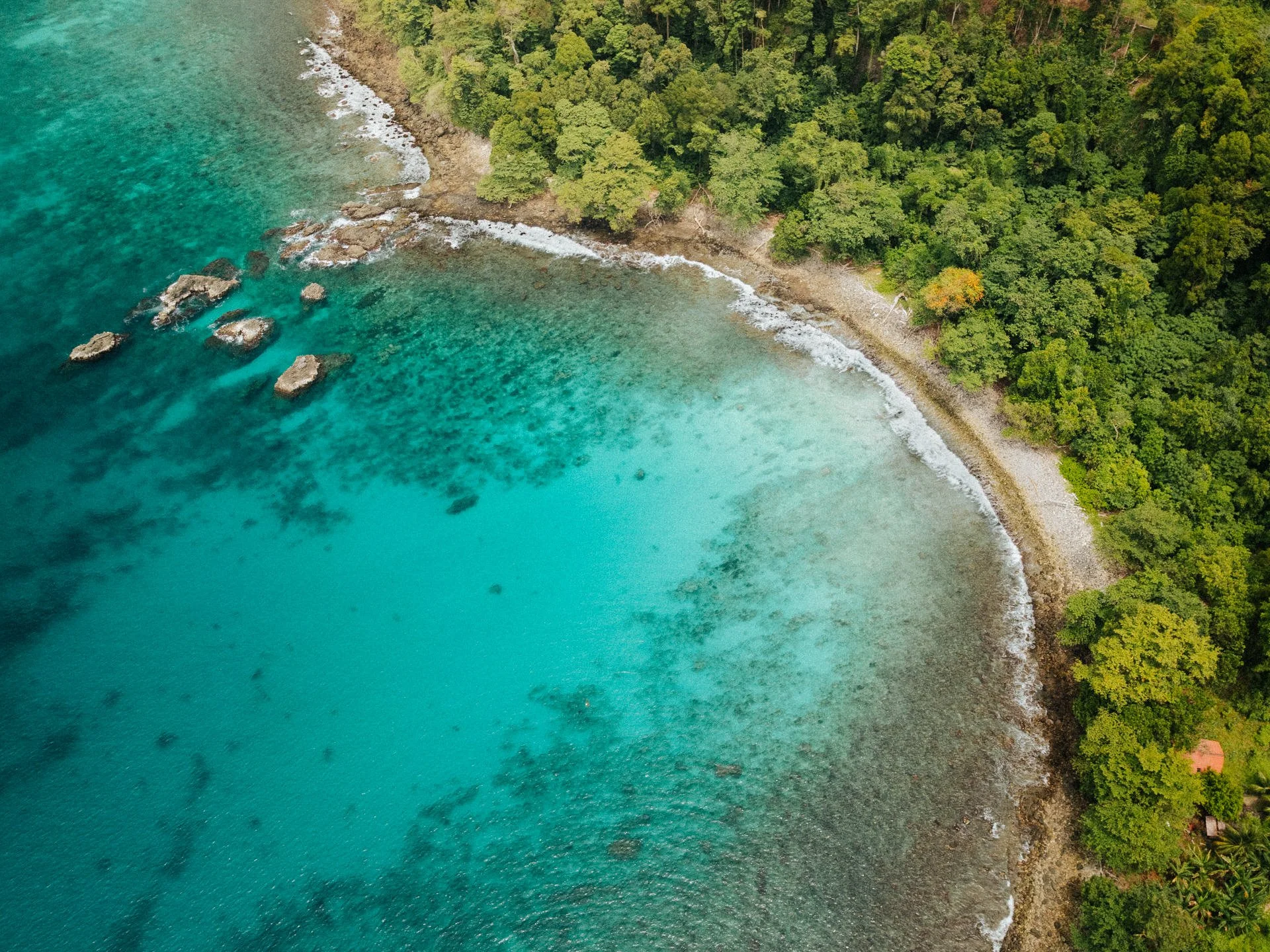 Aerial view of a tropical coastline with turquoise water, rocky shoreline, and dense green forest.