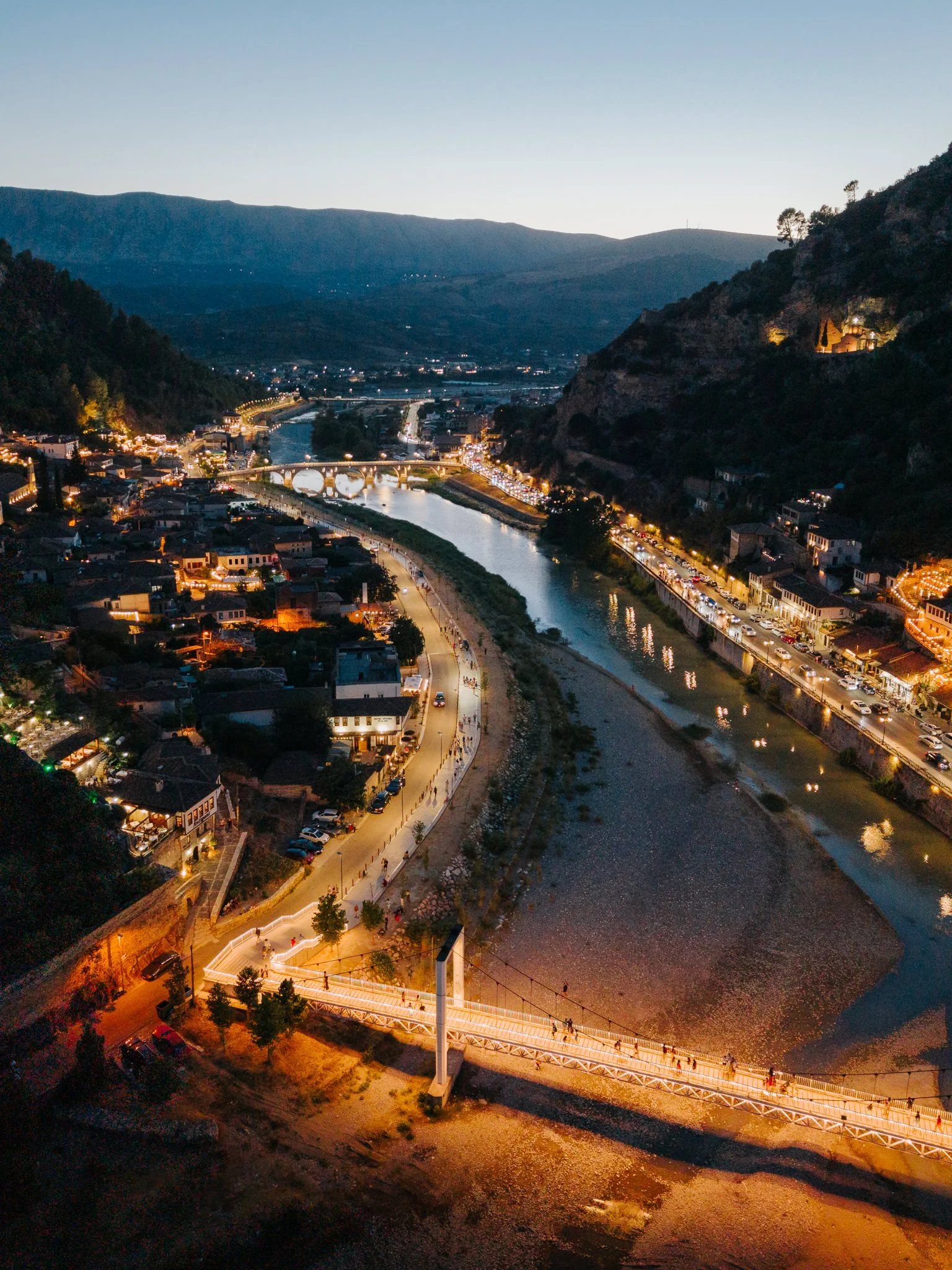 A town by a river with a bridge and mountains in the background, lit by streetlights and buildings at dusk.