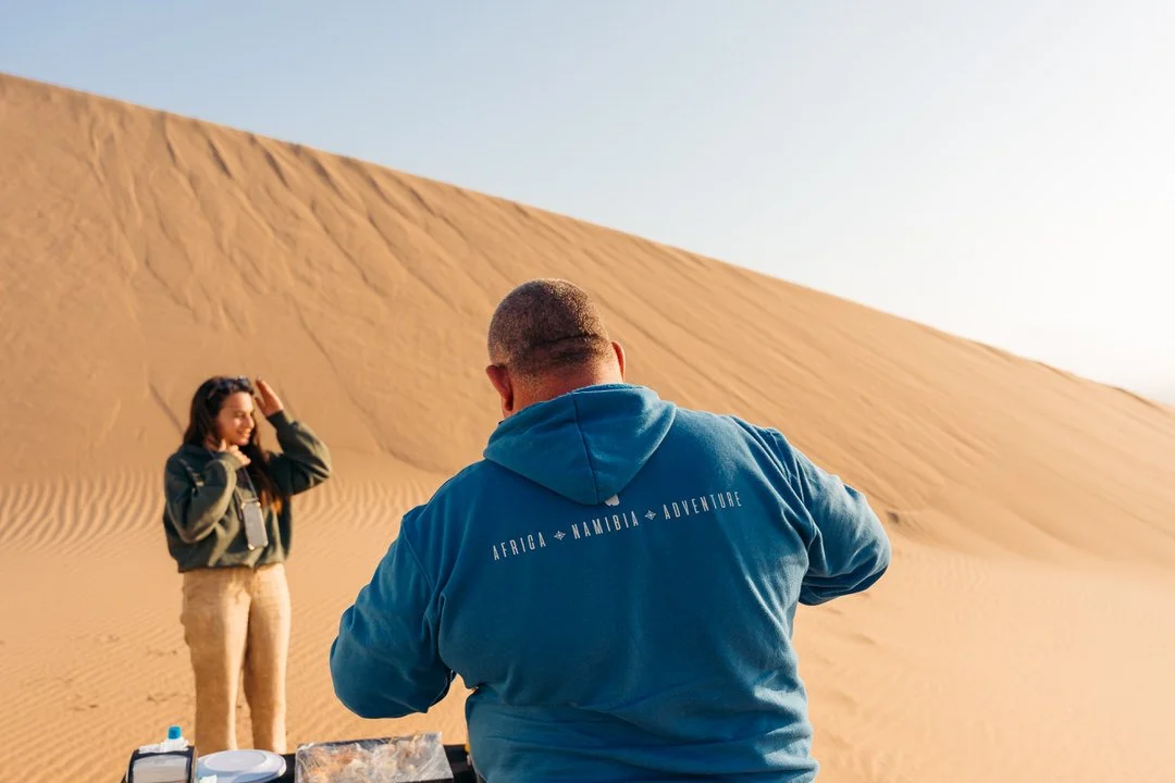A man in a blue hoodie taking a photograph of a woman standing in a desert with large sand dunes.