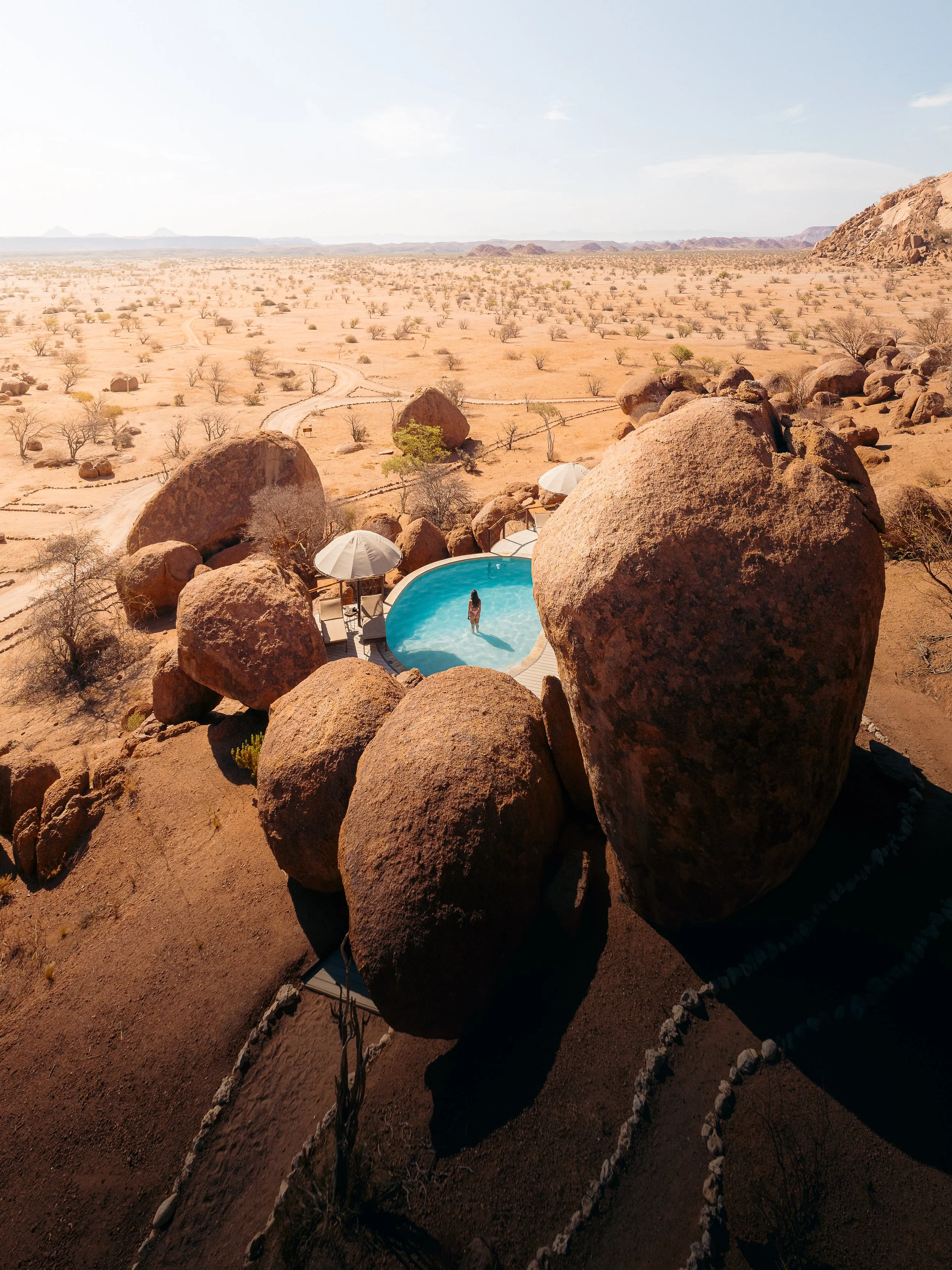 A woman standing in a small, round swimming pool surrounded by large rocks in a desert landscape with sparse vegetation and distant mountains.