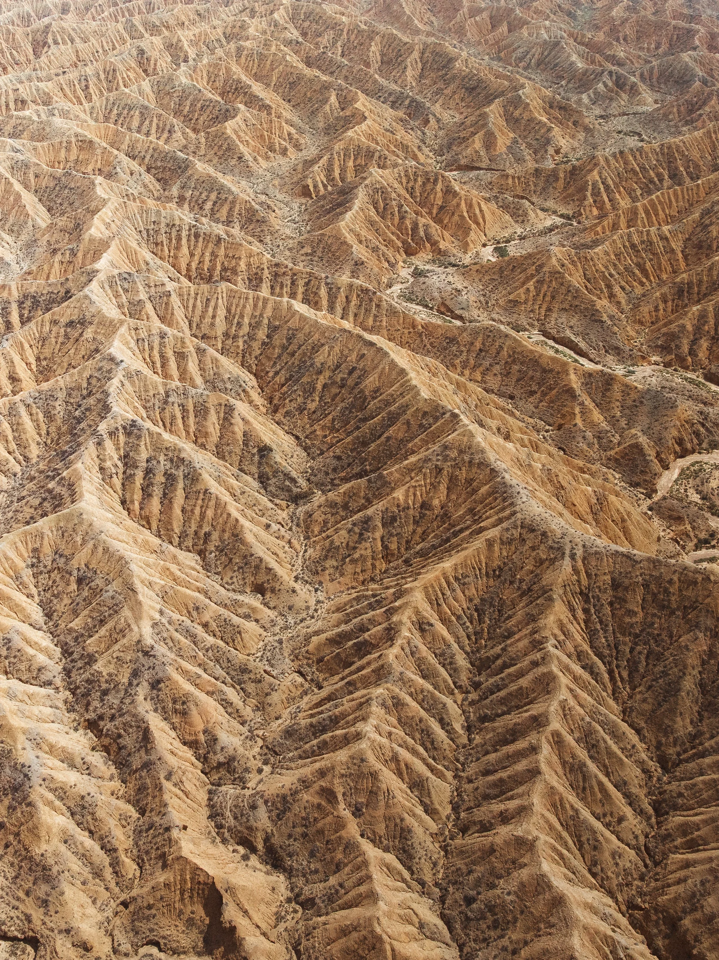 Aerial view of rugged desert mountains with sharp ridges and deep valleys in shades of brown and tan.