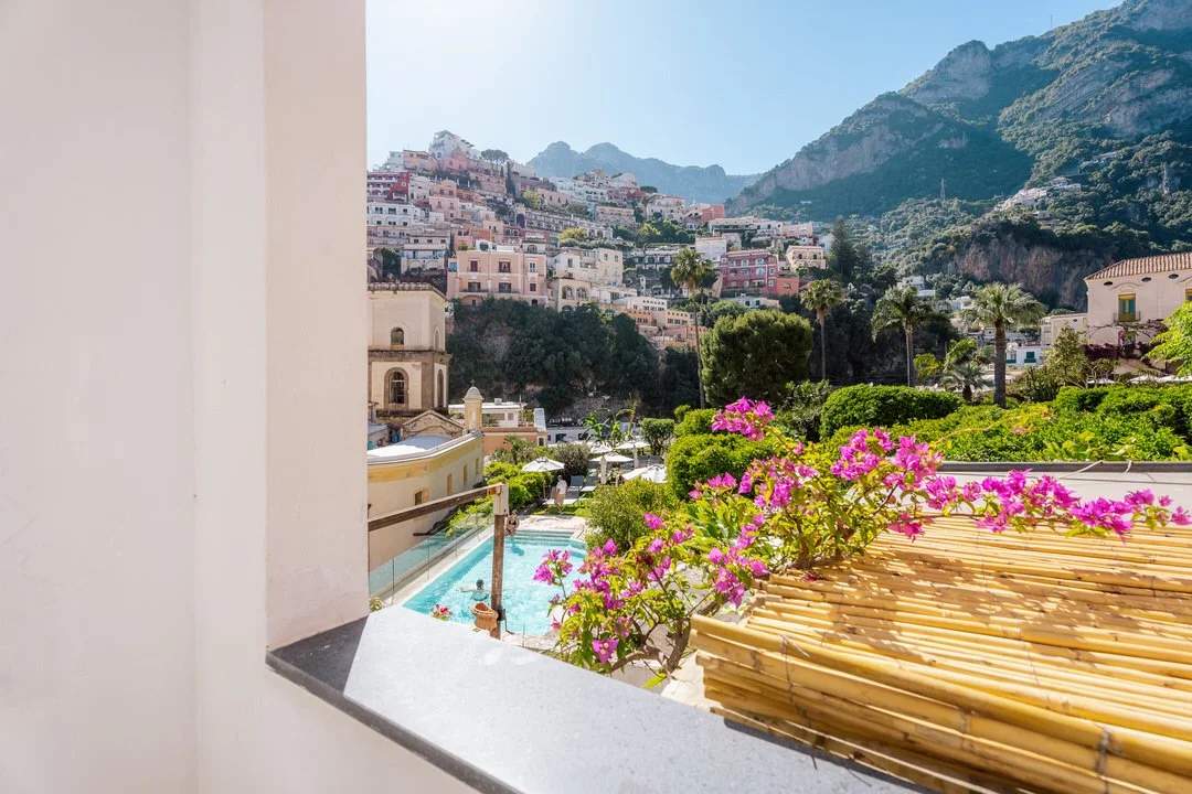 View from a balcony overlooking a hillside town with colorful buildings, lush greenery, pink flowers, a swimming pool, and mountains in the background on a bright sunny day.