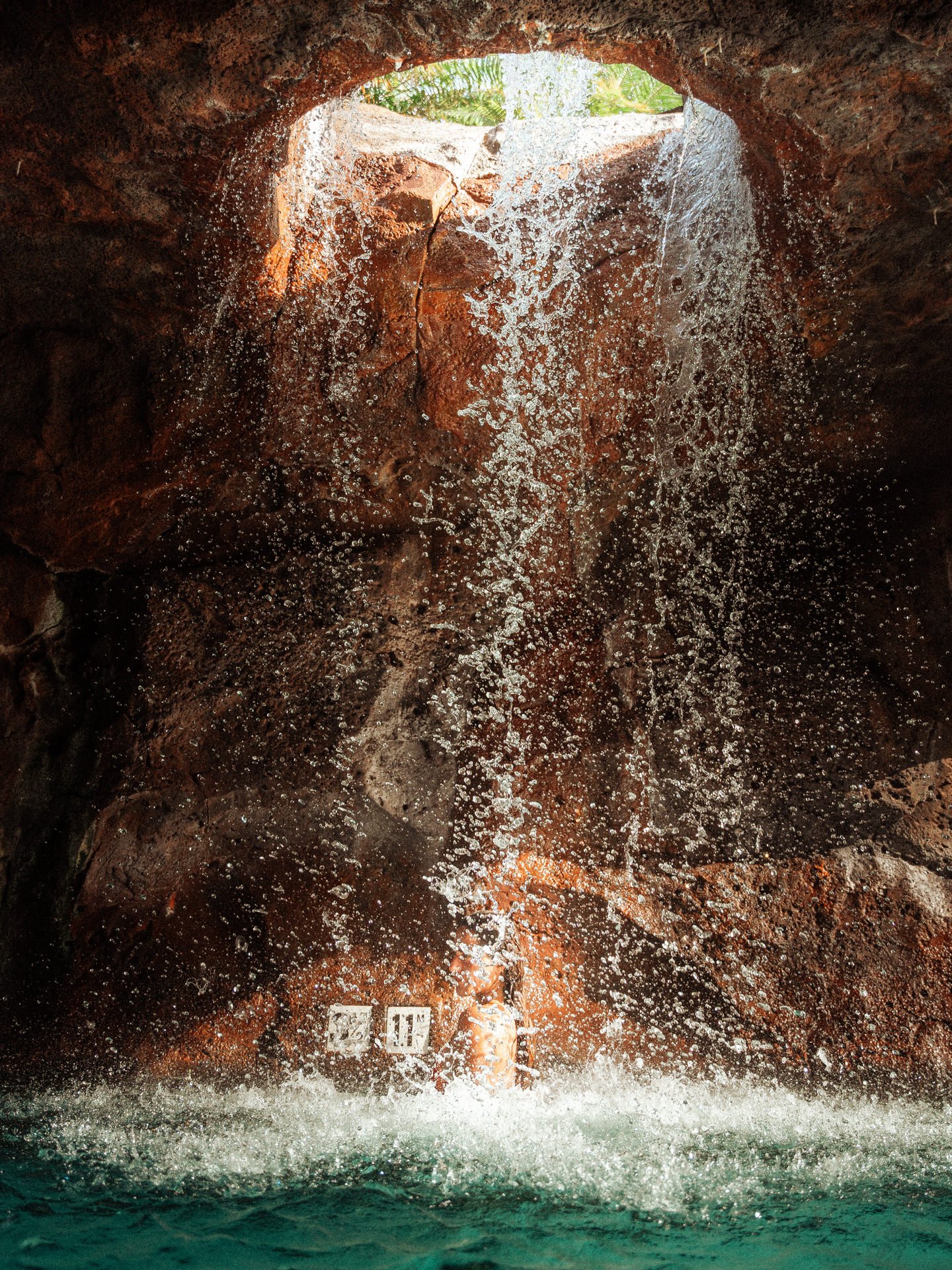 Waterfall flowing over red rocks from an opening in a cave with greenery visible outside.
