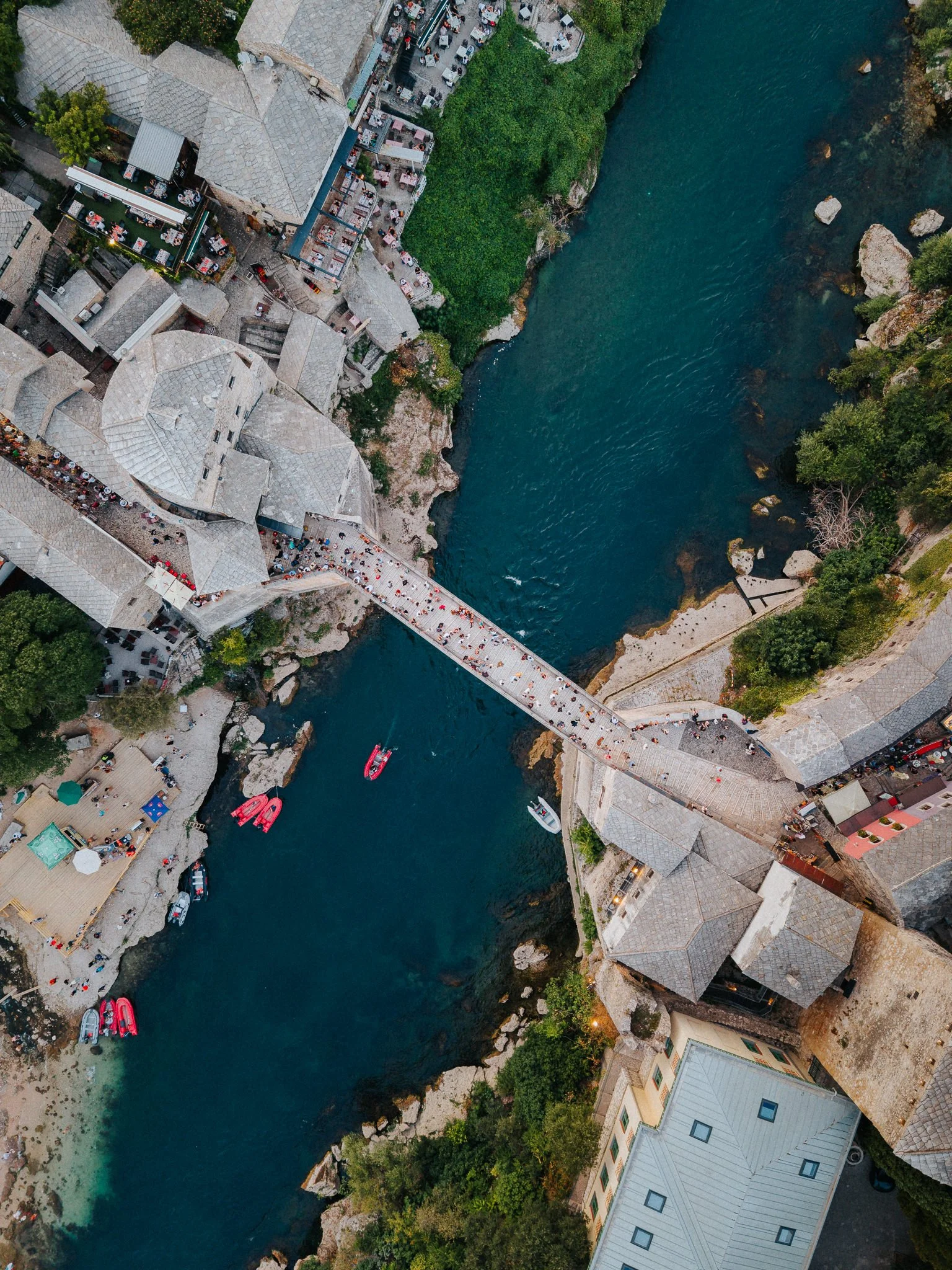 An aerial view of a river with a walkway bridge crossing over it, surrounding buildings with gray roofs, outdoor dining areas, boats on the river, and lush green trees.