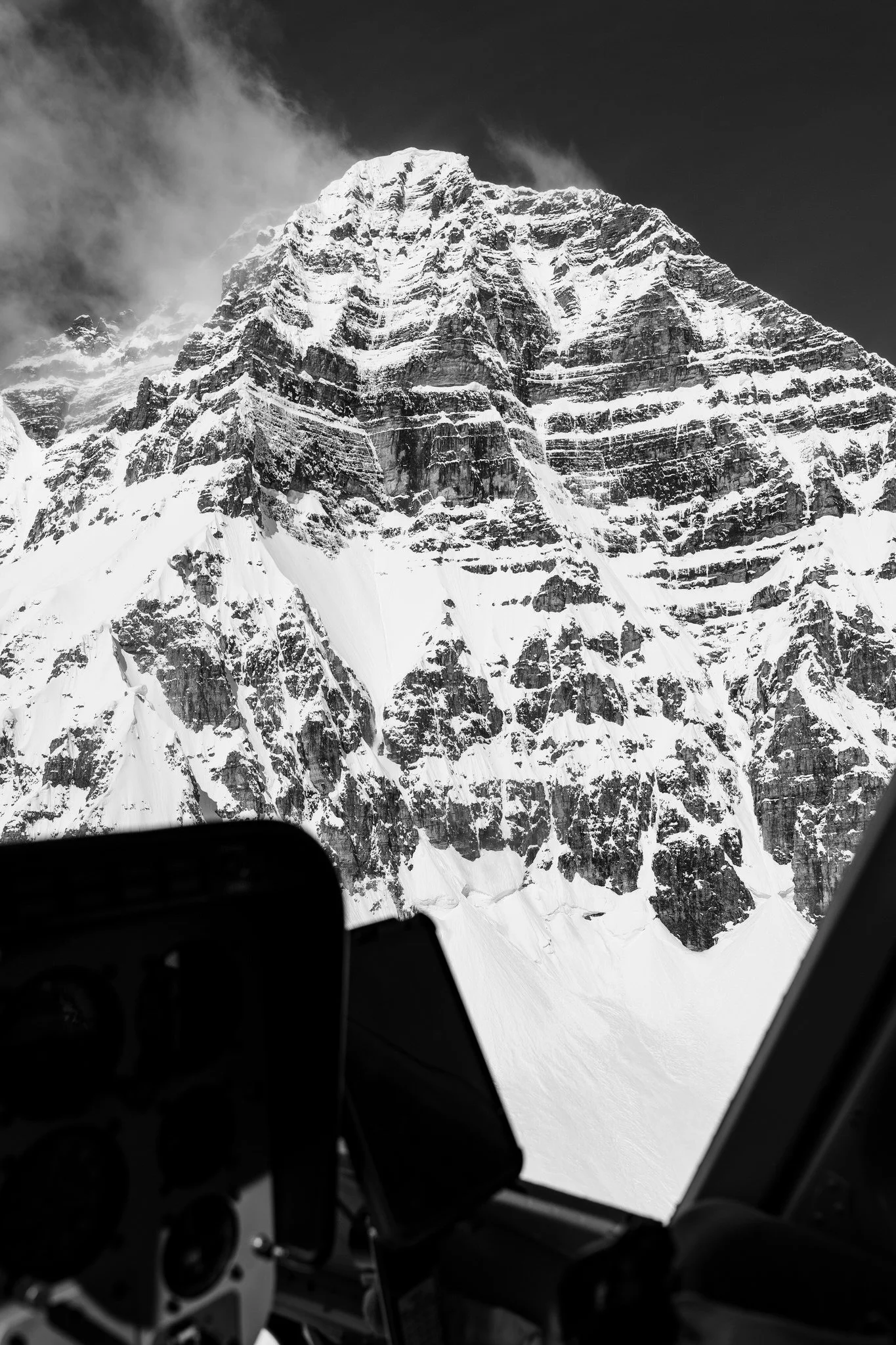 Black and white photo of a snow-covered mountain peak seen from inside an aircraft, with cockpit instruments slightly visible in the foreground.