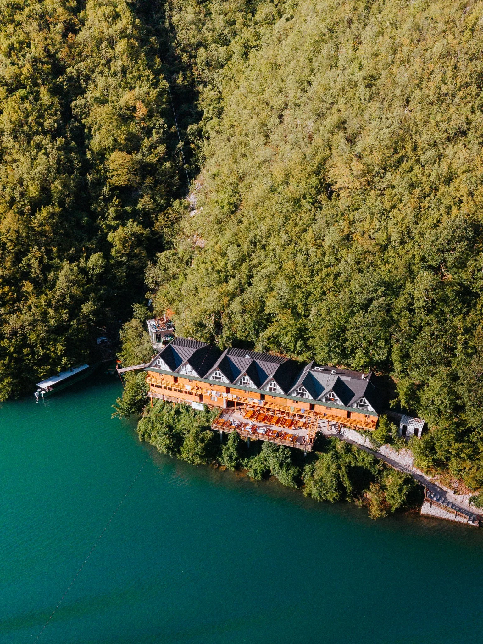Aerial view of a house with a large deck by a green lake, surrounded by dense forest.