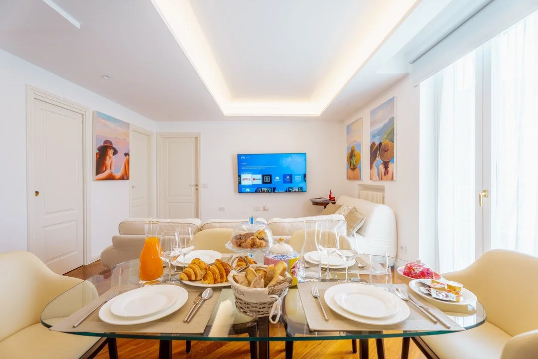 A living room with a glass dining table set for breakfast, featuring plates, glasses, and food, a sofa, wall art, a wall-mounted TV, and large windows with sheer curtains letting in natural light.