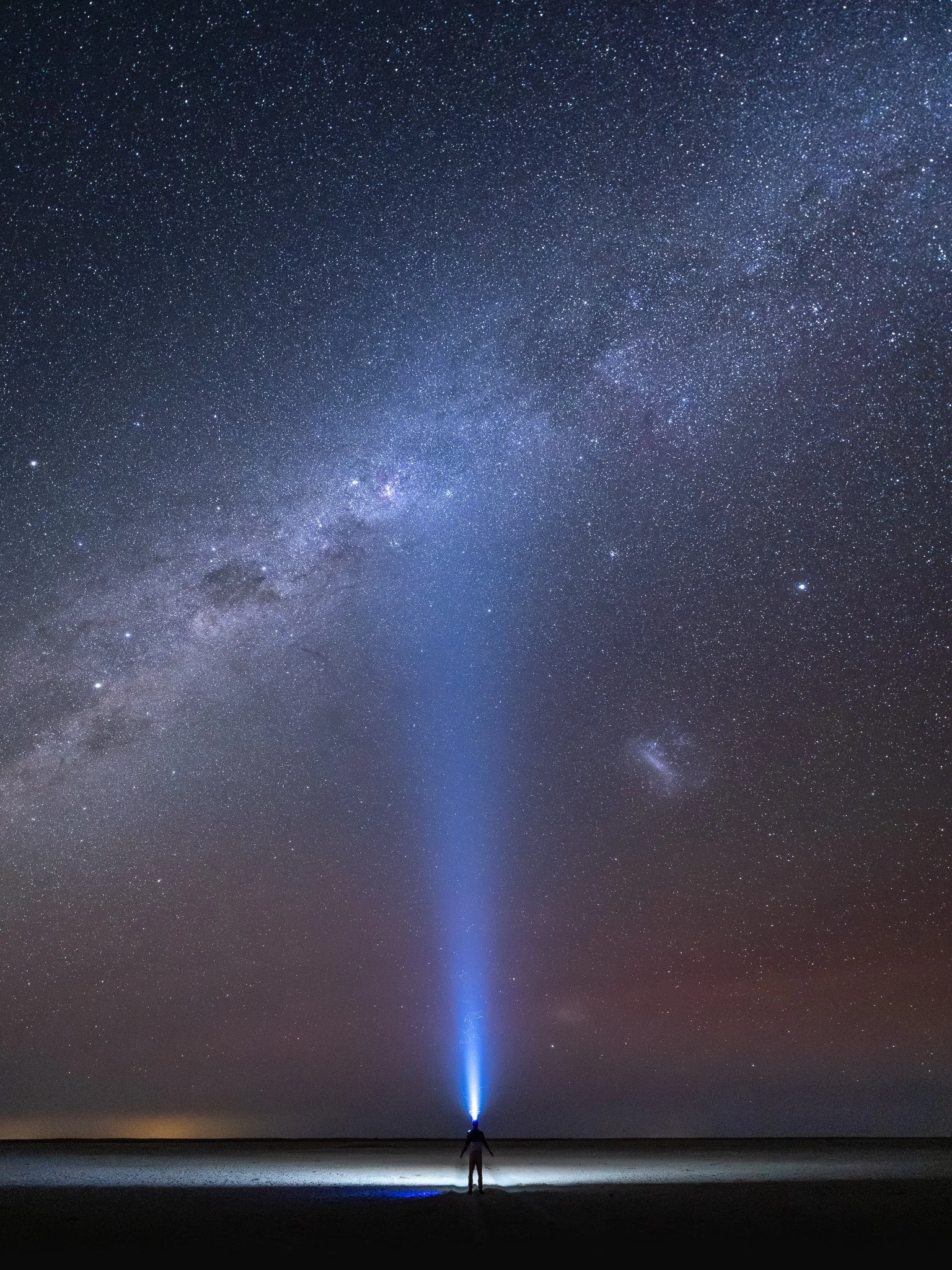 Person standing on a flat surface at night, illuminated by a beam of blue light shooting vertically into the star-filled sky, including the Milky Way Galaxy.