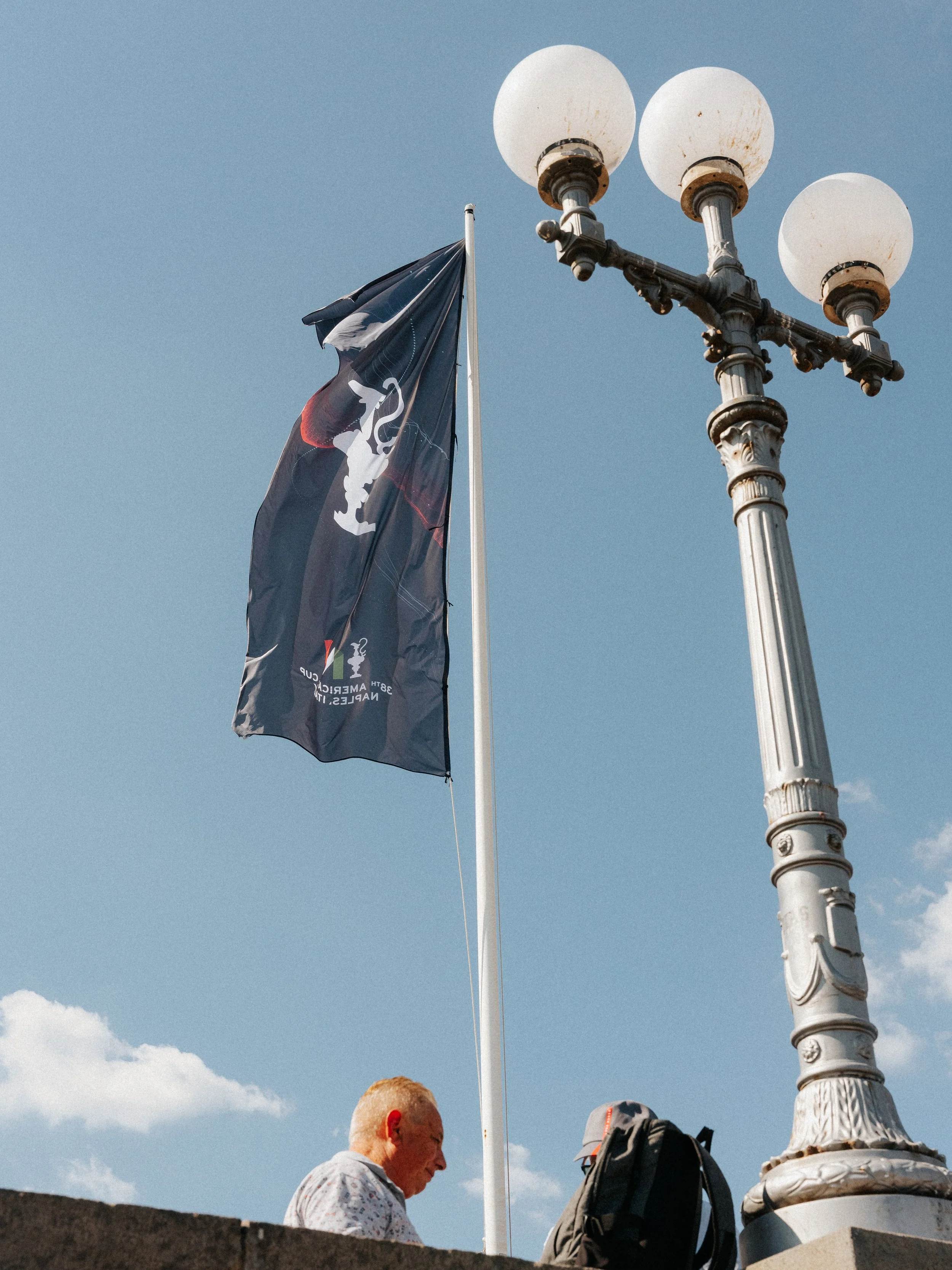 A street lamp with three round, white glass bulbs, a flag on a pole, and two people, one with white hair and a patterned shirt and the other with a backpack, standing near a monument or column against a blue sky with some clouds.