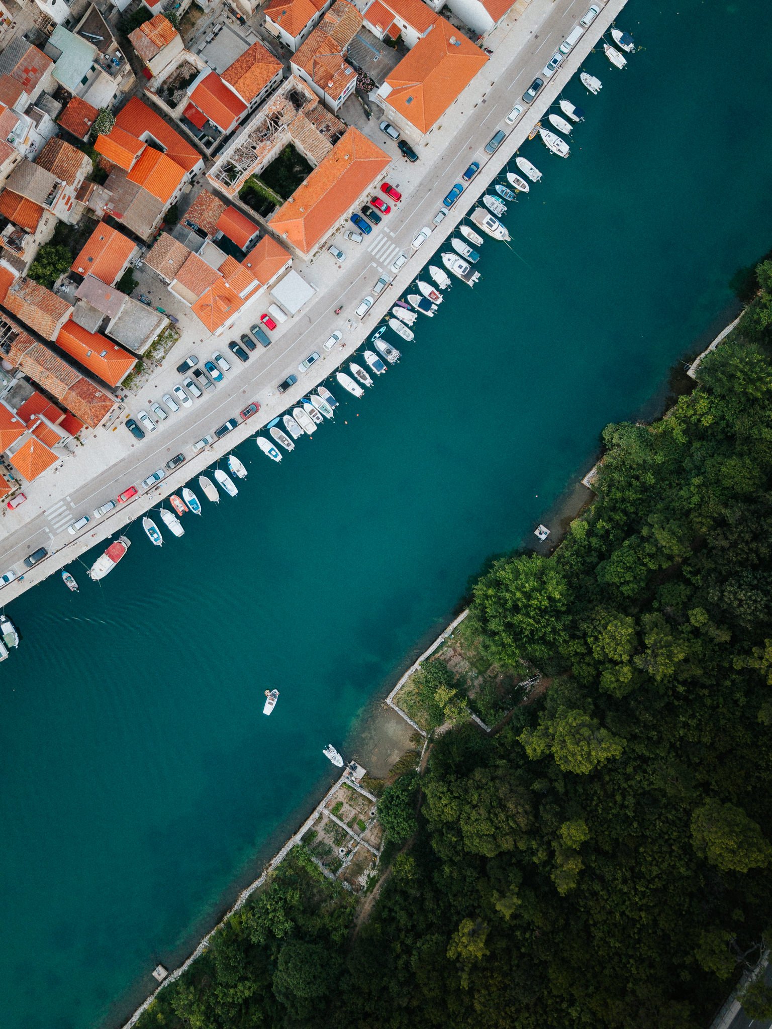 Aerial view of a coastal city with tightly packed buildings, many with red-tiled roofs, along a waterfront with boats docked at the pier, and surrounded by green trees.
