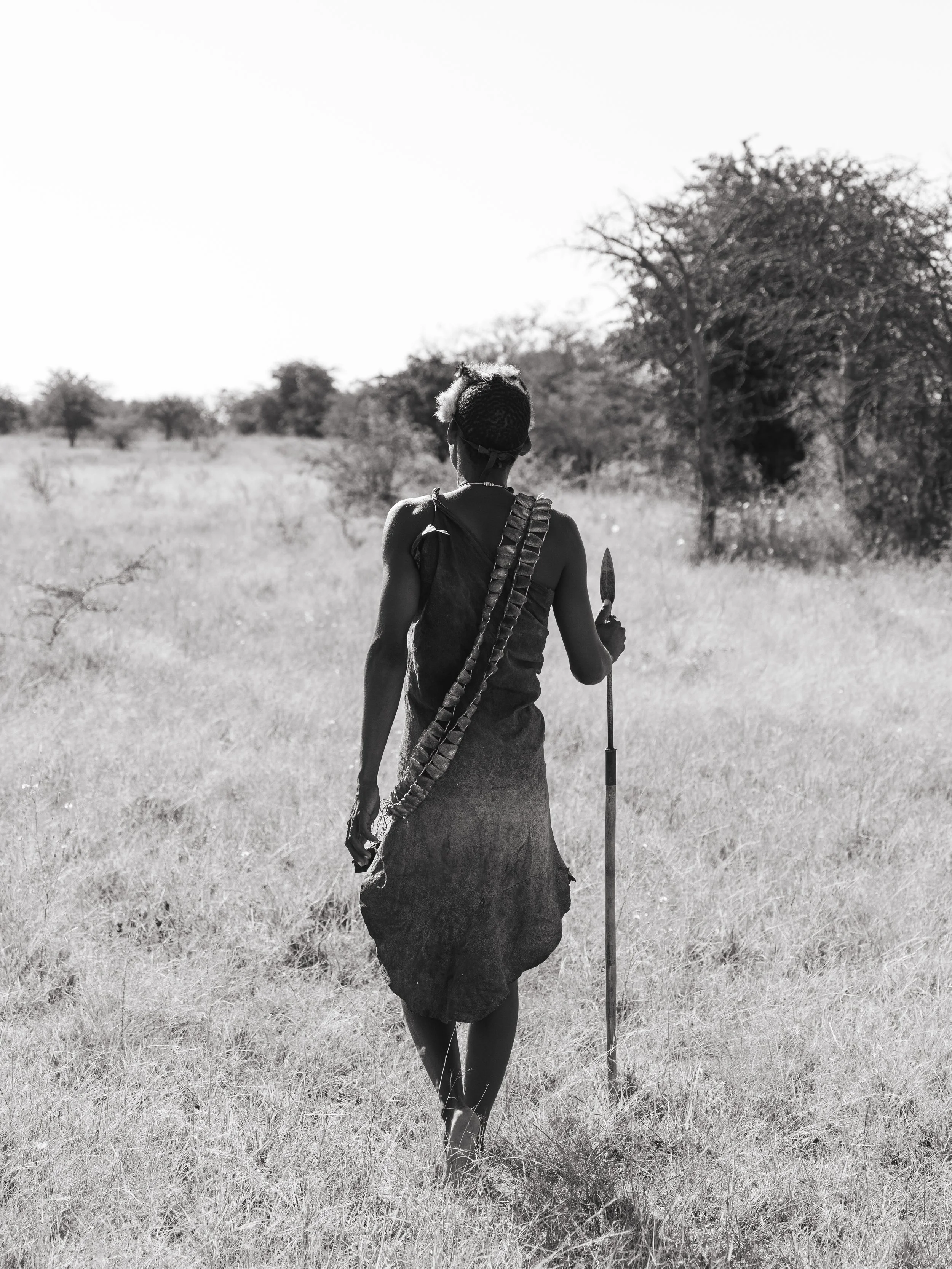 A woman dressed in traditional Maasai attire, walking through a grassy landscape with sparse trees under a clear sky.