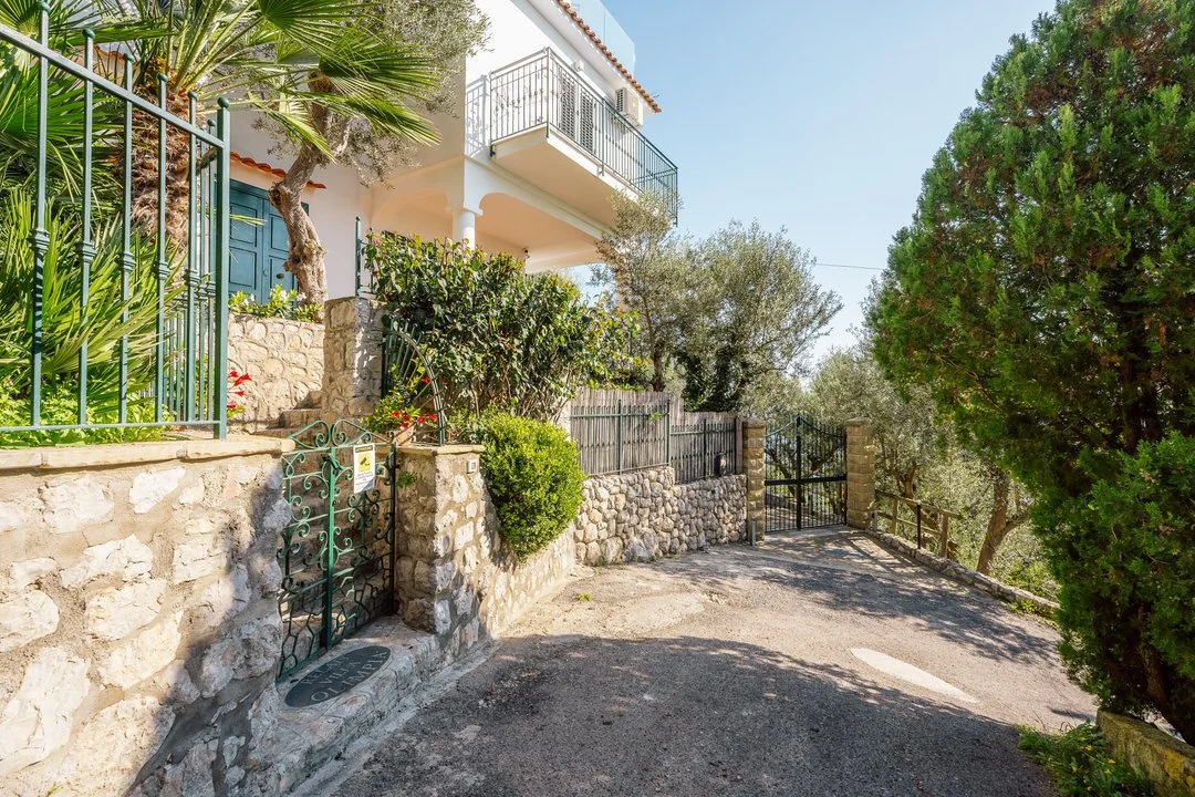 A residential house with a white exterior and balconies, surrounded by greenery, a stone wall with a small decorative gate, and a driveway leading to a gated entrance on a sunny day.