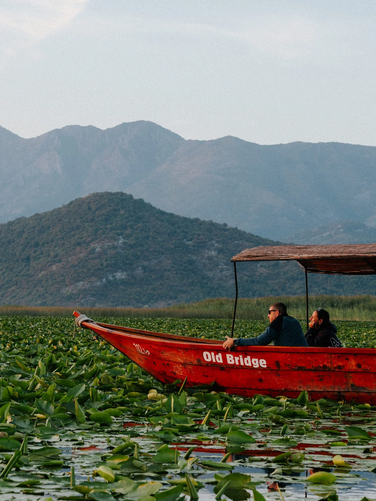 A red boat labeled 'Old Bridge' floating on a lily pad-covered lake with two people sitting inside, and mountain ranges in the background.