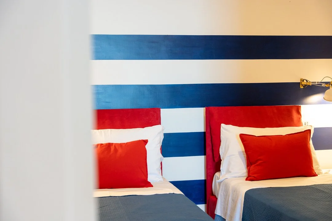 Two hotel beds with red headboards, red pillows, and white and grey bedding, against a striped blue and white wall, with a wall-mounted lamp on the right side.