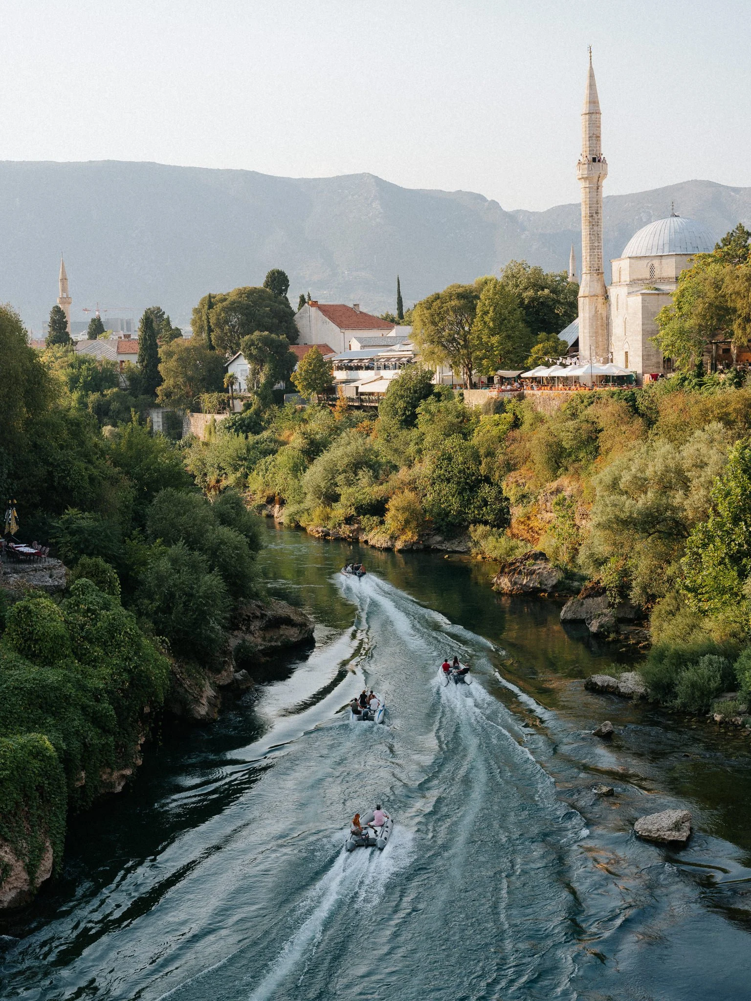 Boats with people on a river surrounded by lush green trees and buildings with minarets, with mountains in the background.