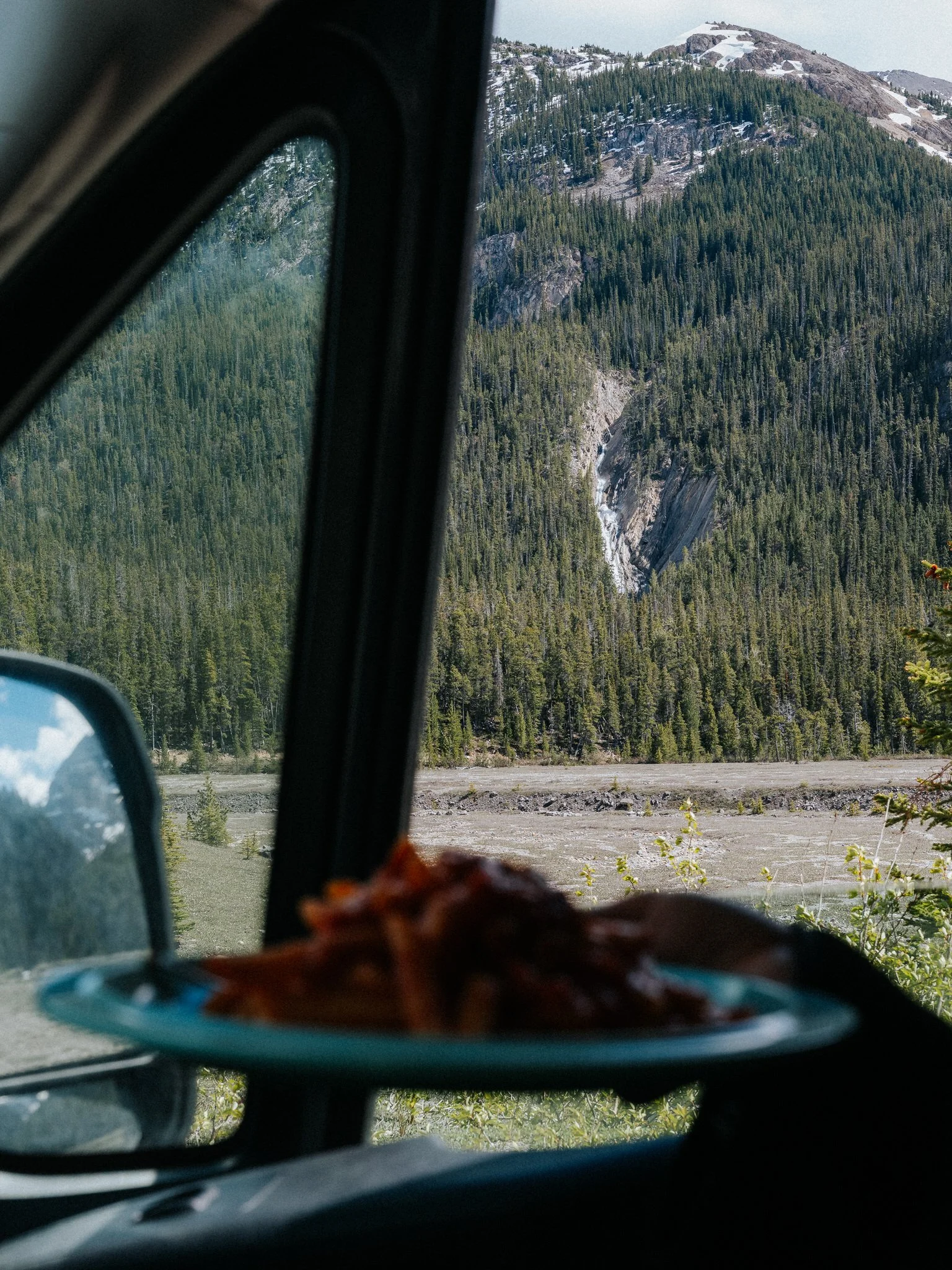A view of a mountainous landscape taken from inside a vehicle, showing a snow-capped mountain, dense evergreen forest, and a waterfall in the distance.